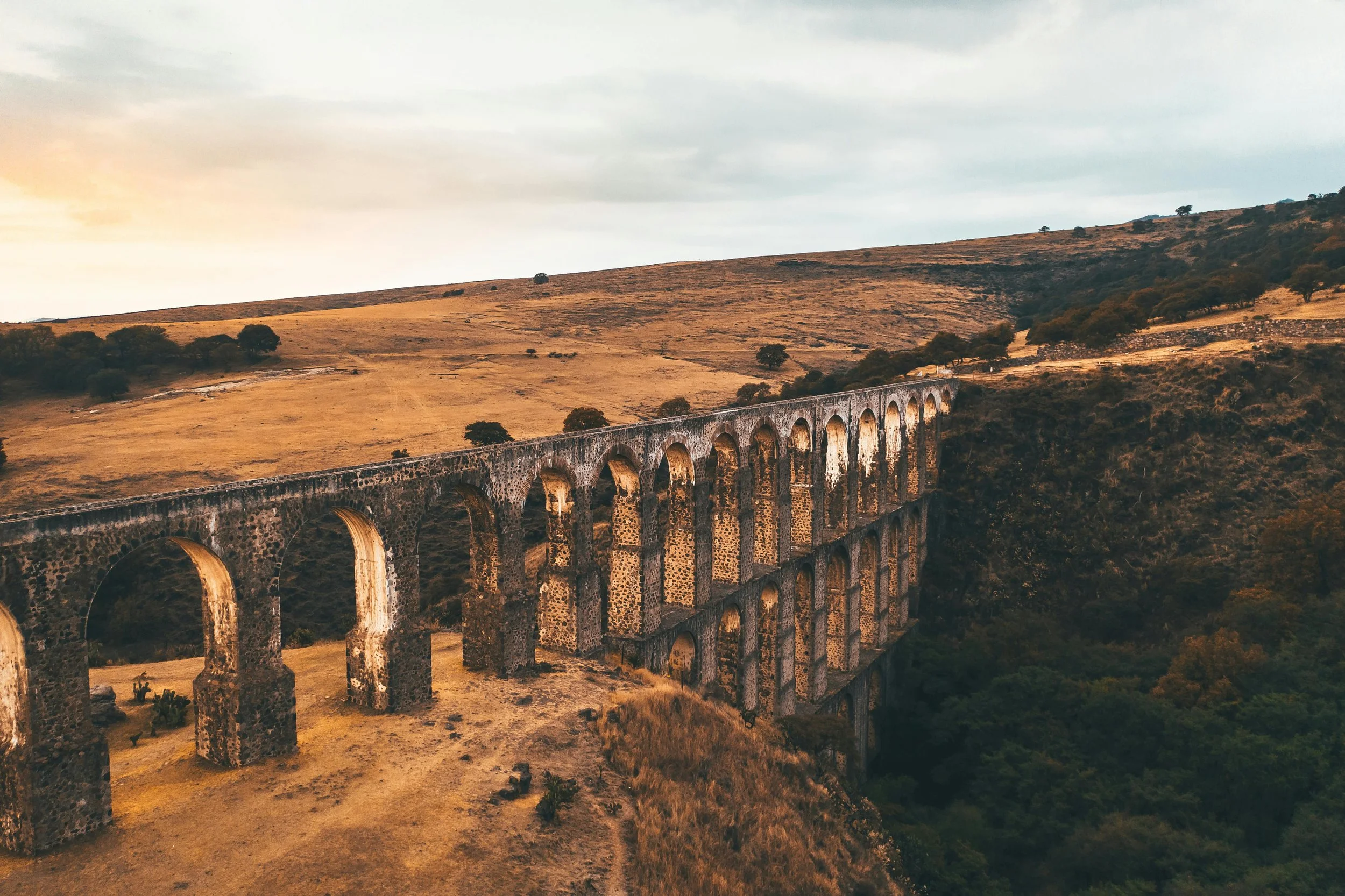 An old stone aqueduct spanning across a dry, grassy hillside with scattered trees under a cloudy sky during sunset.