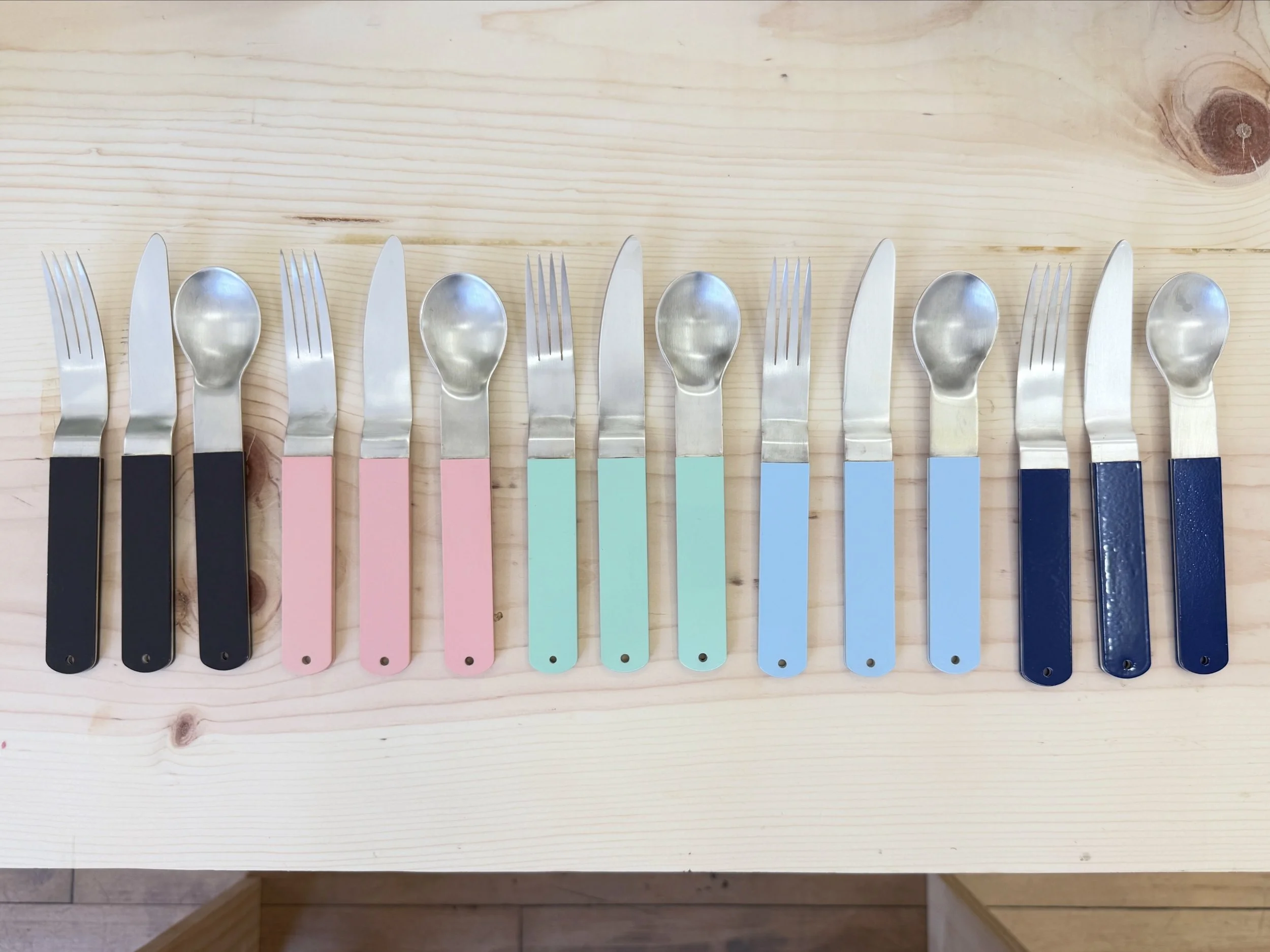 Set of metal utensils with dark brown, pink, mint green, and navy blue handles on a wooden surface.
