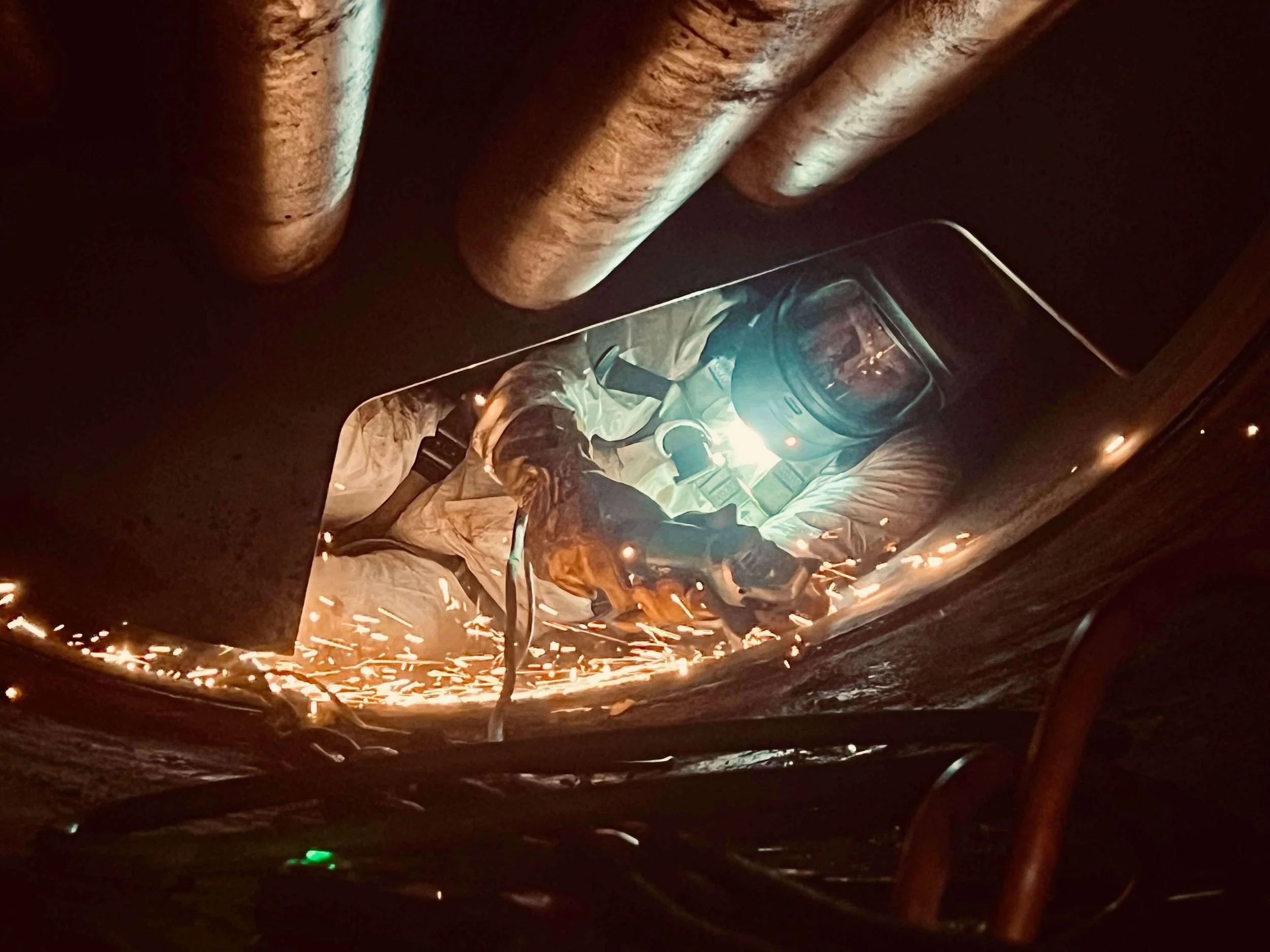 A welder wearing protective gear and a helmet welding metal with bright sparks flying in an industrial setting.