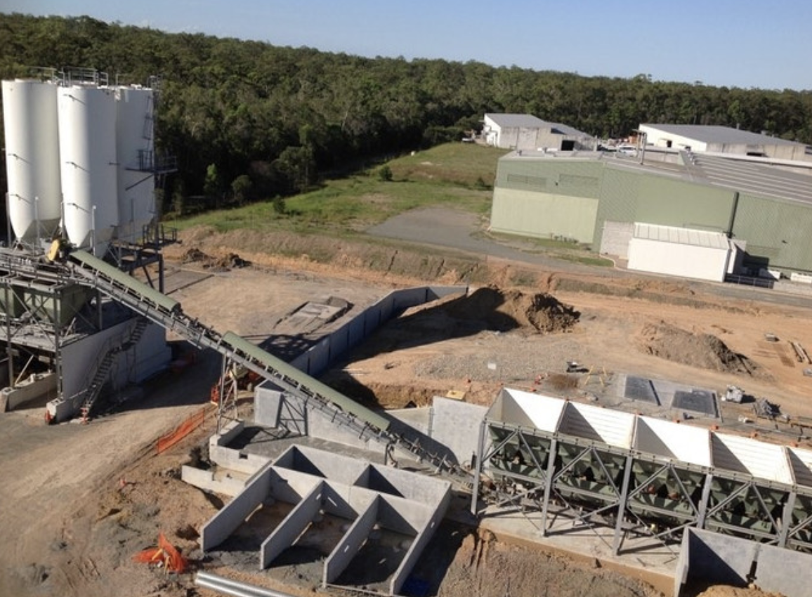 An industrial construction site with large storage tanks, metal structures, and ongoing groundwork, surrounded by green forested hills.