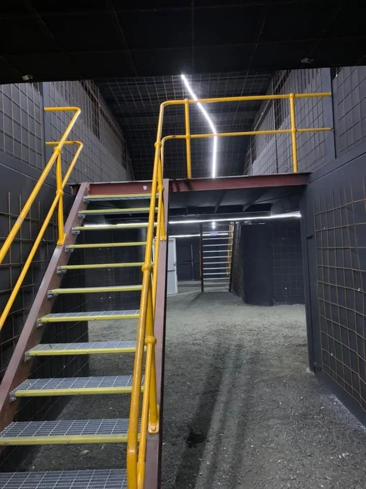 Industrial stairway with yellow guardrails leading to upper level in a dark interior space with black tiled walls.