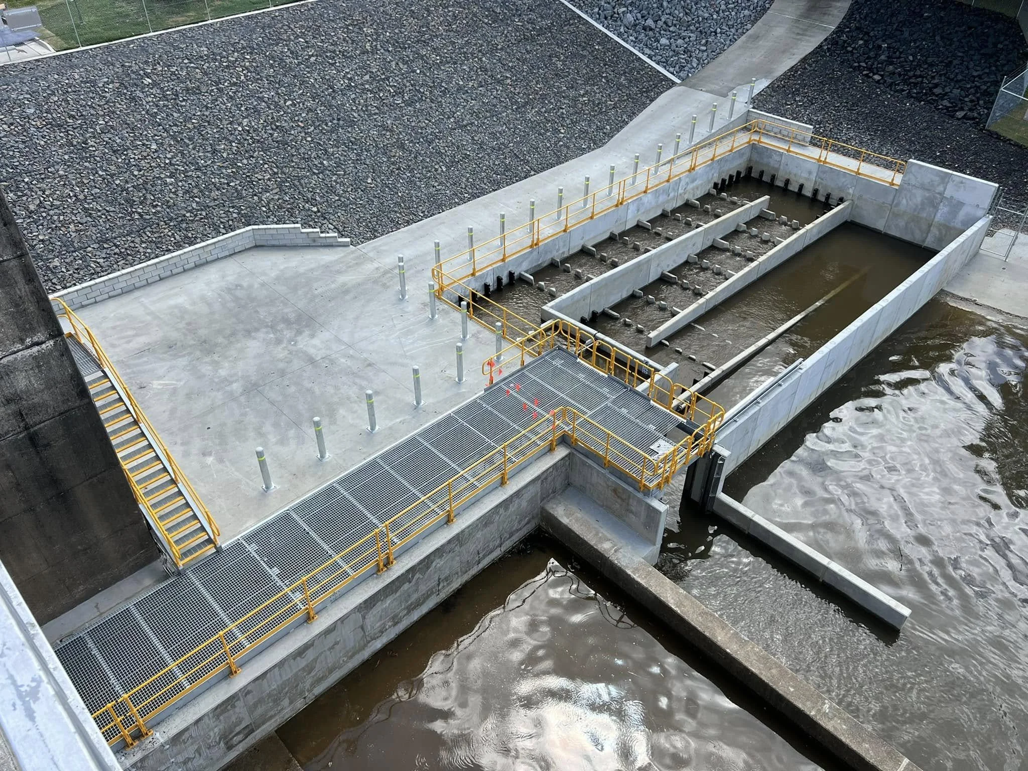 A water treatment facility with concrete basins, yellow railings, and staircases near a body of water.