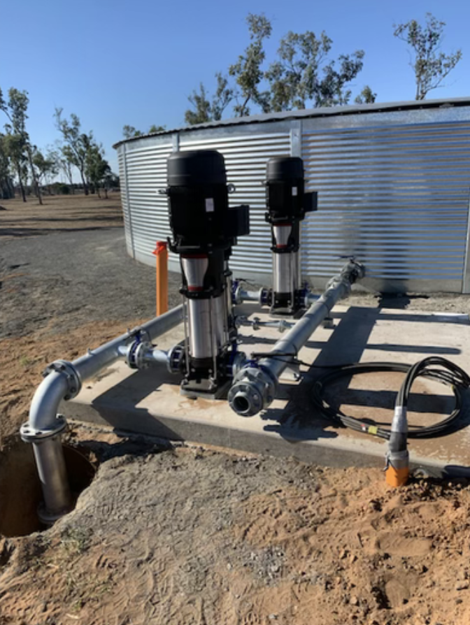 Two vertical black and silver pumps connected to a metal pipeline system outside near a large metal tank in an open, dry area with sparse trees.