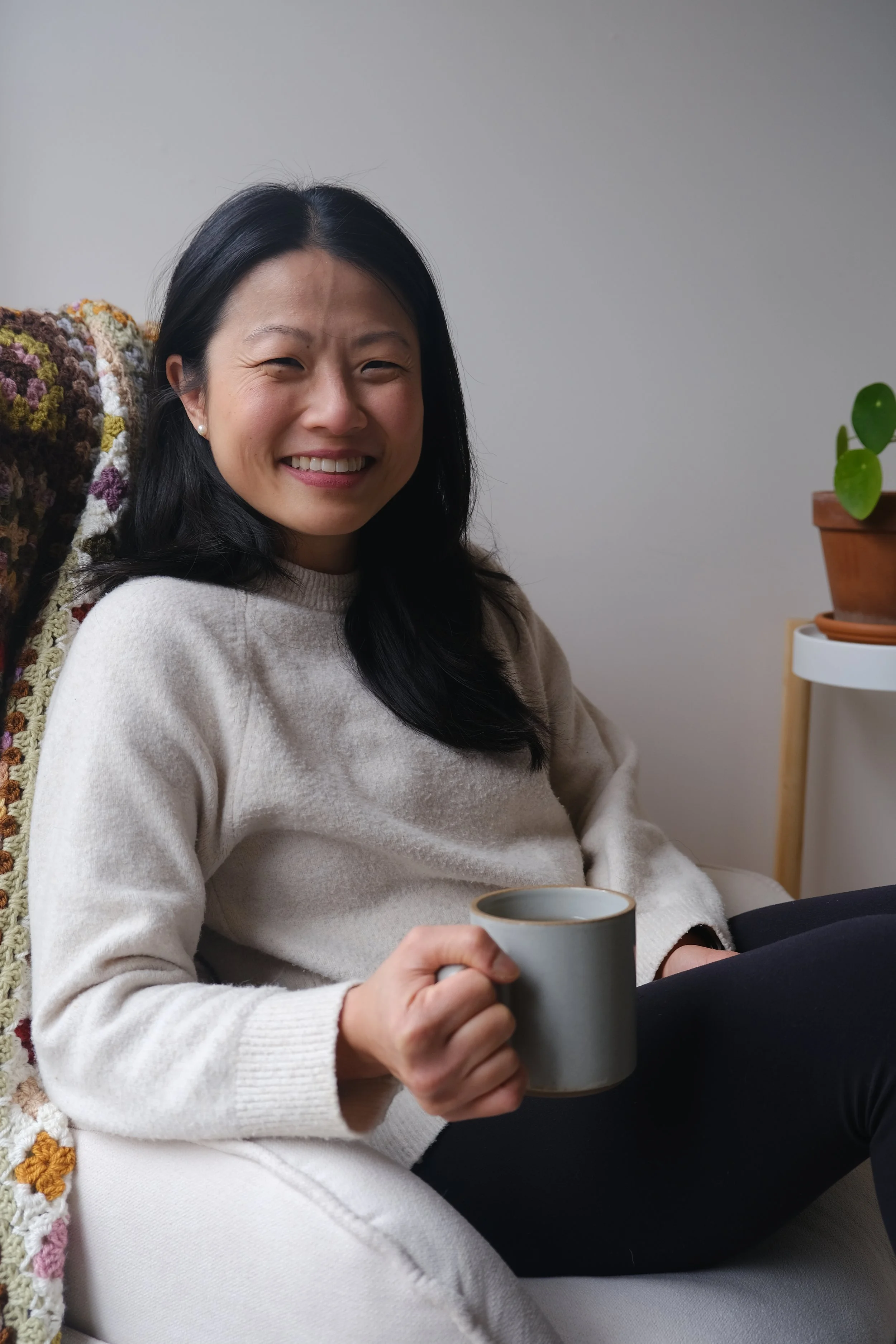 A smiling woman with black hair sitting on a white chair, holding a grey mug, wearing a beige sweater, with a crocheted blanket over her shoulder. There is a white side table with a potted plant in the background.