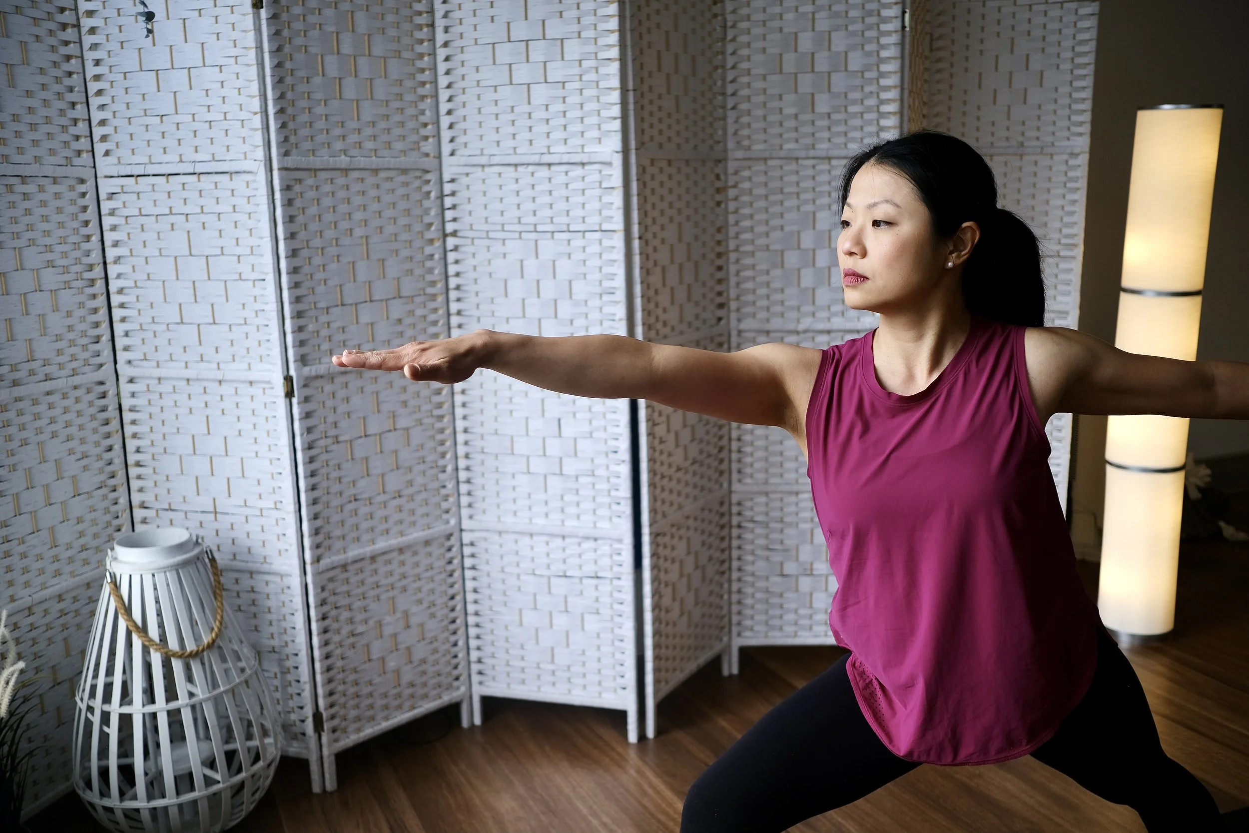 Woman in pink sleeveless top and black leggings practicing yoga or stretching indoors, with a white wicker room divider and floor lamp in the background.