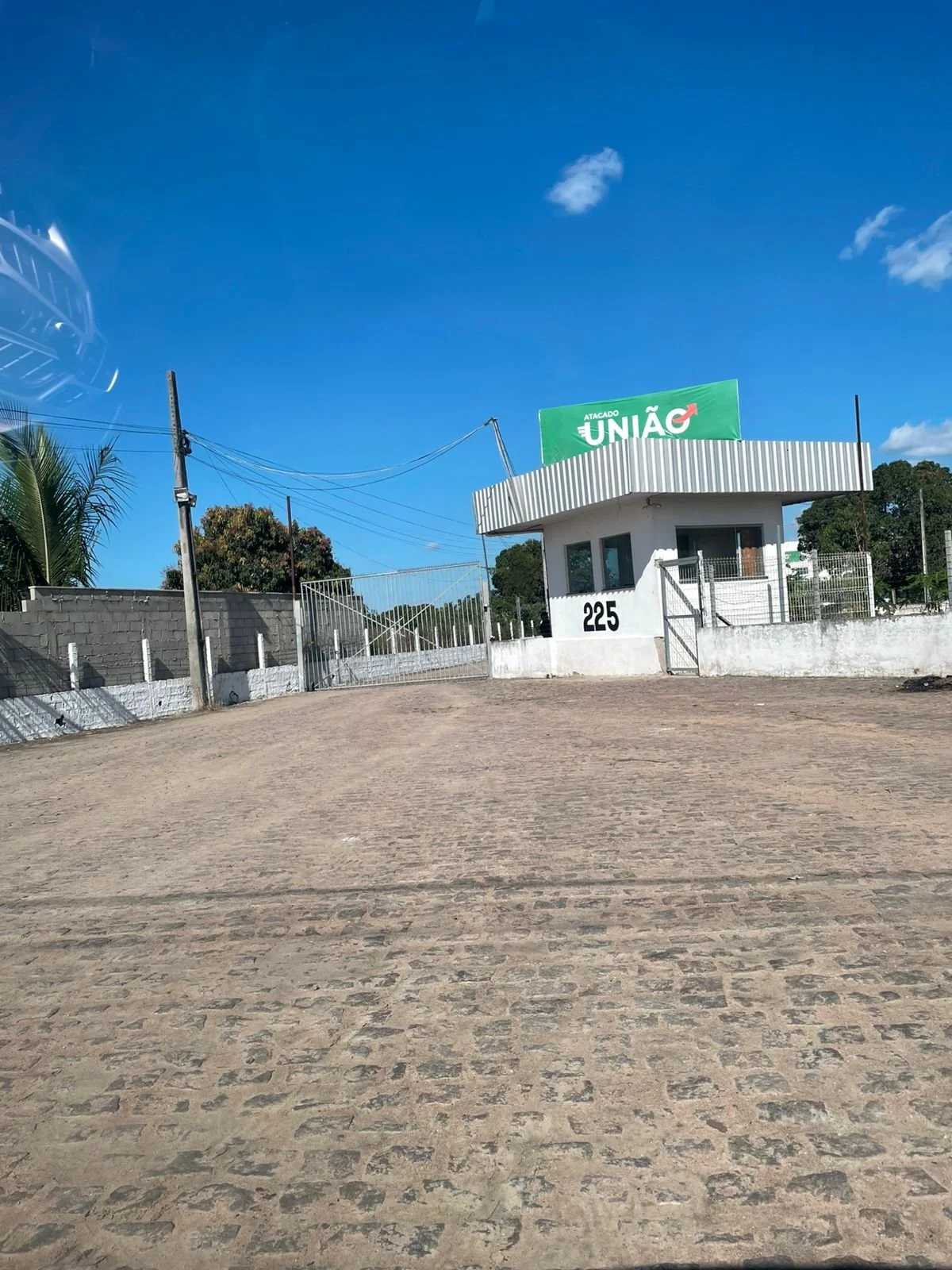 Entrada de uma propriedade com muro branco, portão de ferro e um guarda de cabine de controle com placa de número 225, sob um céu azul com poucas nuvens, e publicidade de campanha política com a palavra 'União'.