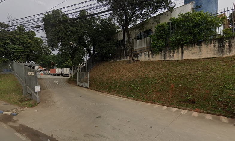 Entrada de rua com portão de ferro aberto, com uma placa de aviso, em uma área urbana com árvores e cerca na lateral, e um declive de gramado ao lado da rua.