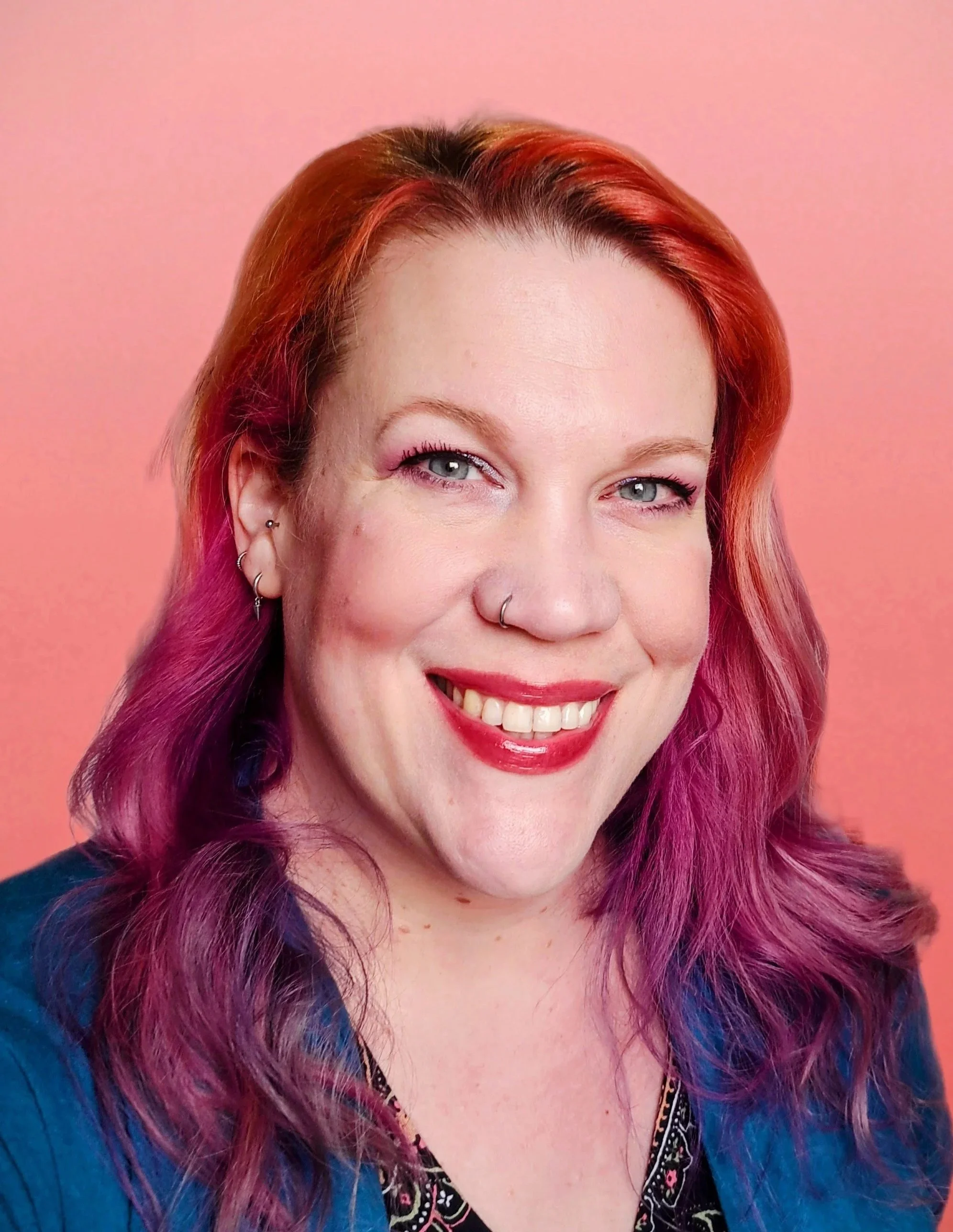Smiling woman with blue eyes, rainbow-coloured hair, wearing earrings and a colourful top, against a pink background.