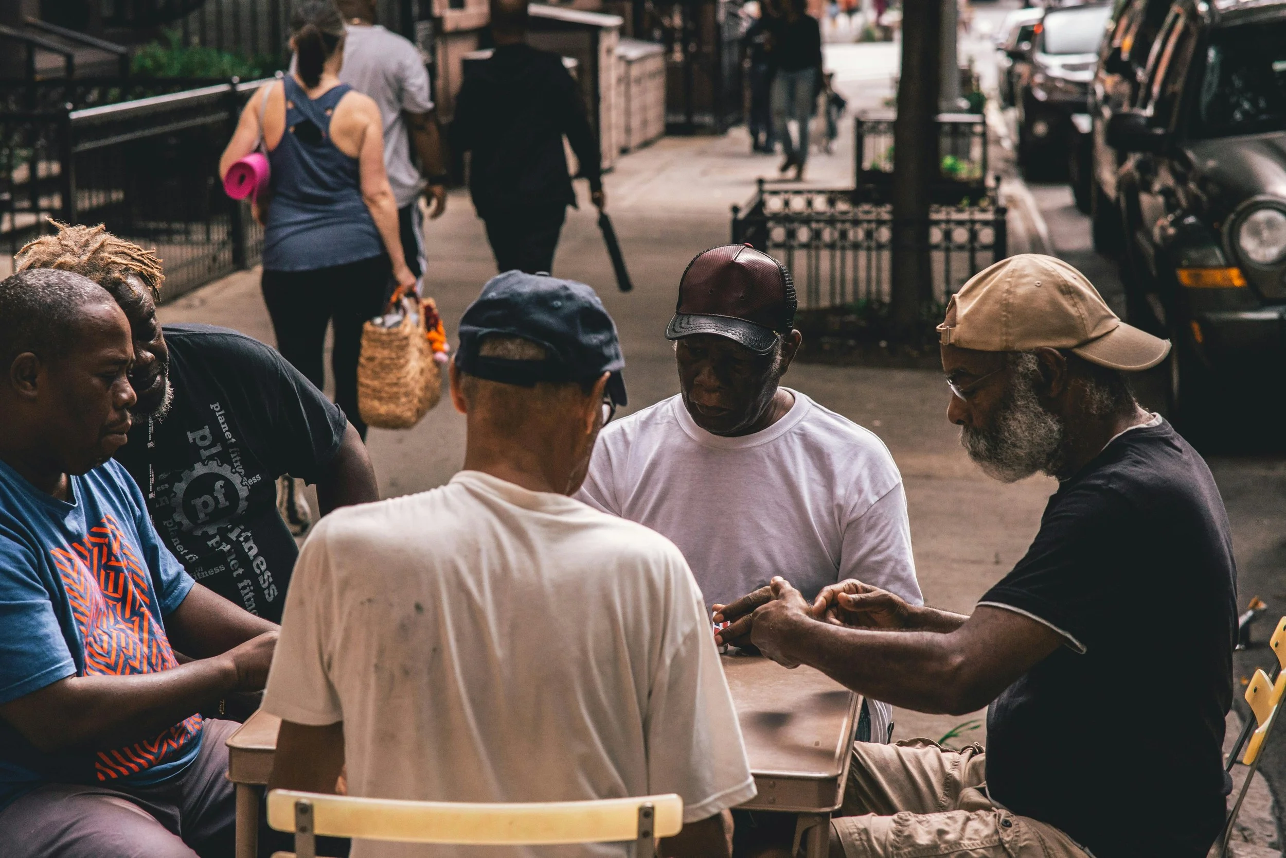 Four men playing a game of cards at a street table on a city sidewalk as pedestrians walk by.