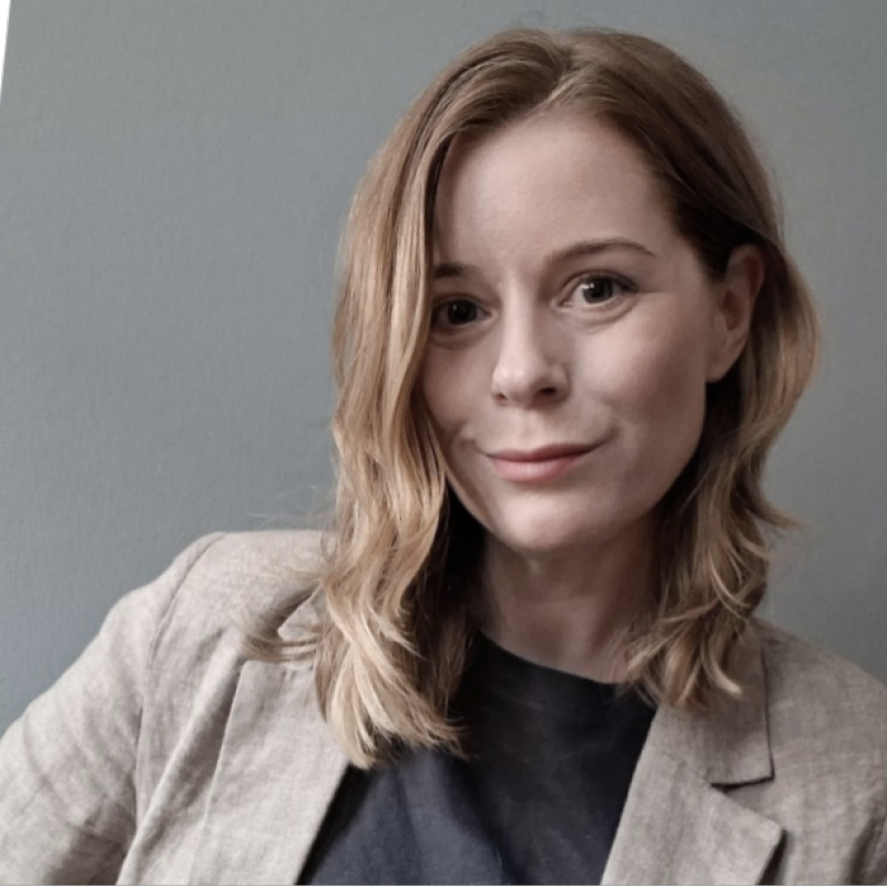 A smiling woman with light brown hair in a black top and beige blazer