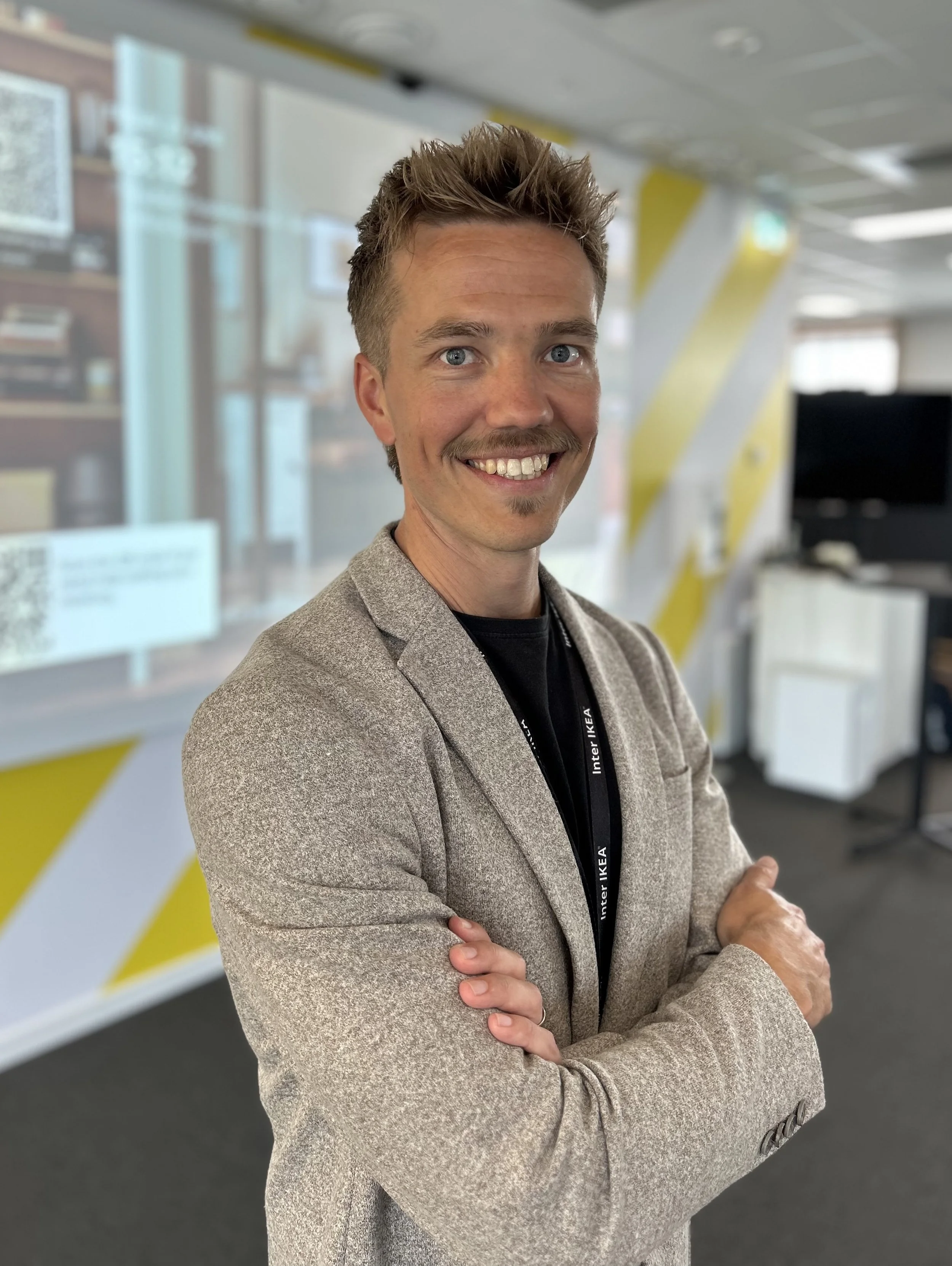 A man standing in an office with crossed arms, smiling, with a blurred background showing office furniture and colorful wall design.
