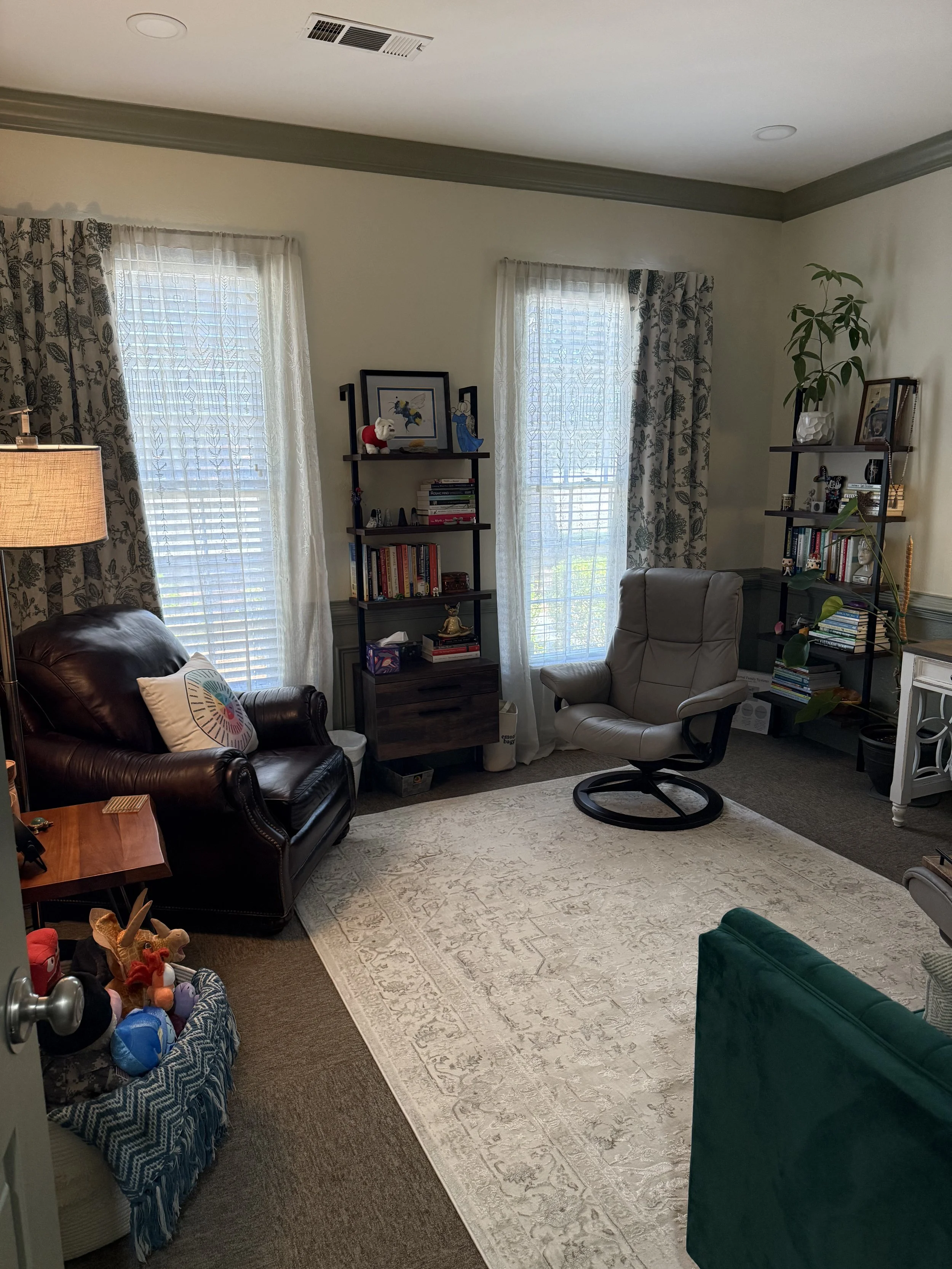 A cozy living room with two large windows covered with lace and floral curtains, a beige armchair, a dark leather armchair with a colorful pillow, a green velvet sofa, a white rug, a wooden side table, bookshelves with books and decorations, toys in a basket, a standing lamp, and a small white desk with a plant on it.