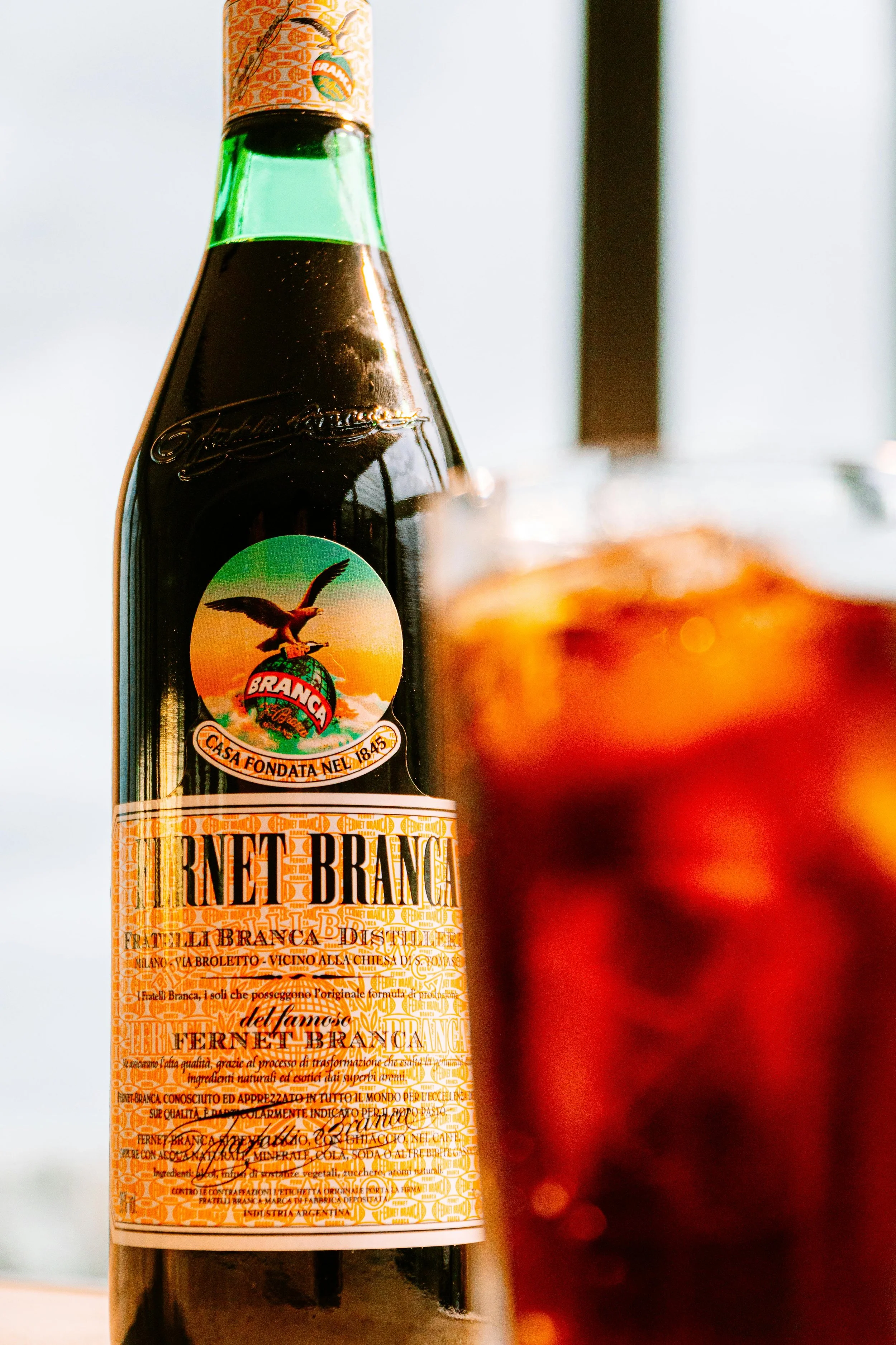 Botella de Fernet Branca en primer plano con un vaso con bebida oscura y hielo en primer plano, parcialmente borroso.
