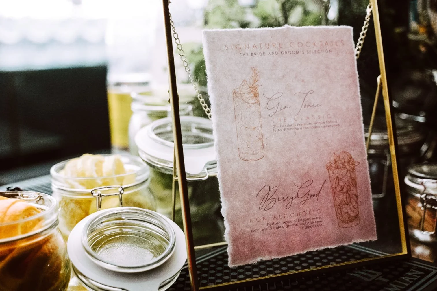 Menú en un soporte dorado, con opciones de cócteles sin alcohol y con alcohol, decorado con ilustraciones y texto en italiano y inglés, rodeado de frascos con ingredientes o toppings en una mesa de madera.