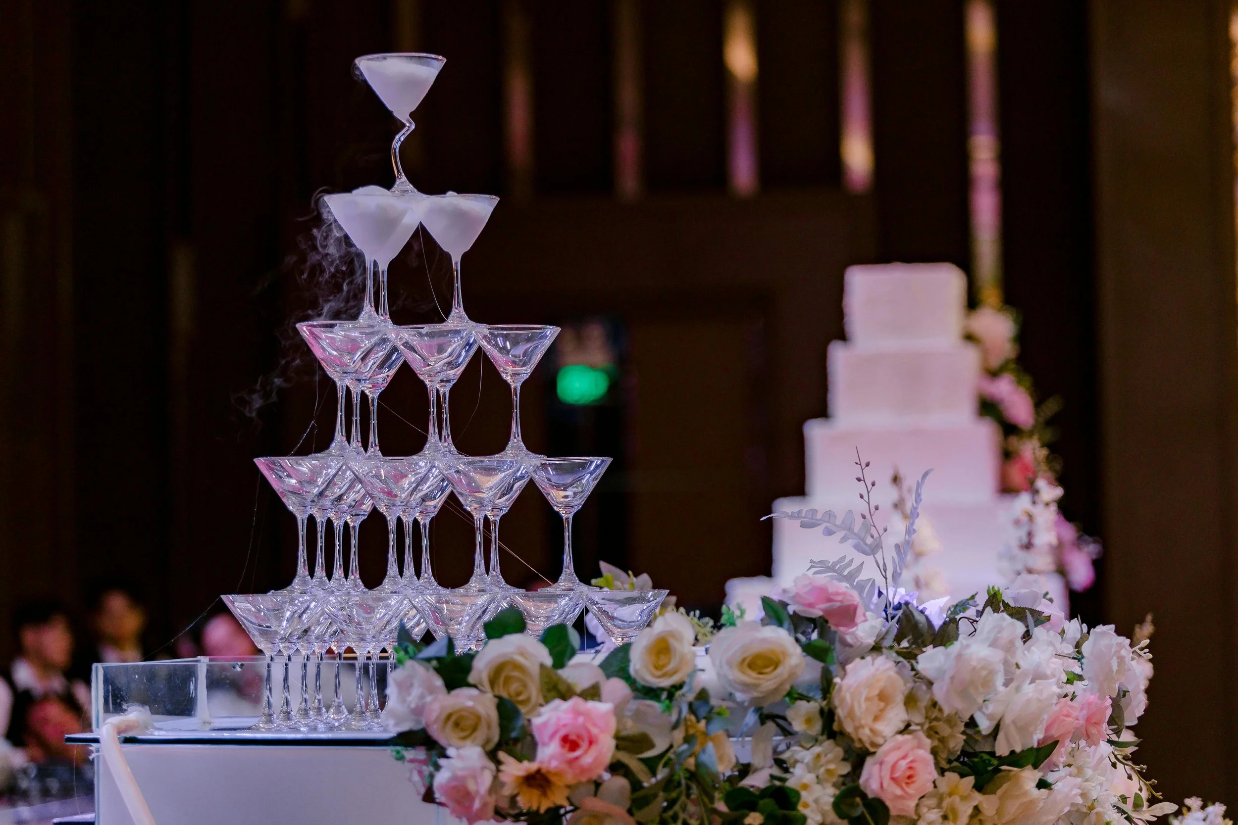 Torre de copas de champán en forma de pirámide con humo y un pastel de bodas decorado con flores en el fondo.