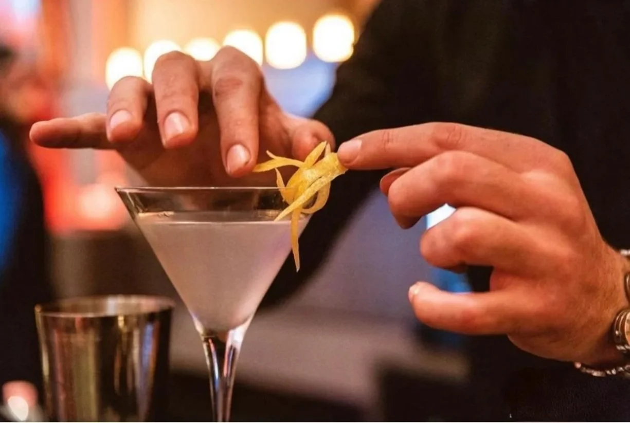 Un cóctel en una copa de Martini decorado con un twist de limón, en un bar con luces suaves y un fondo desenfocado.