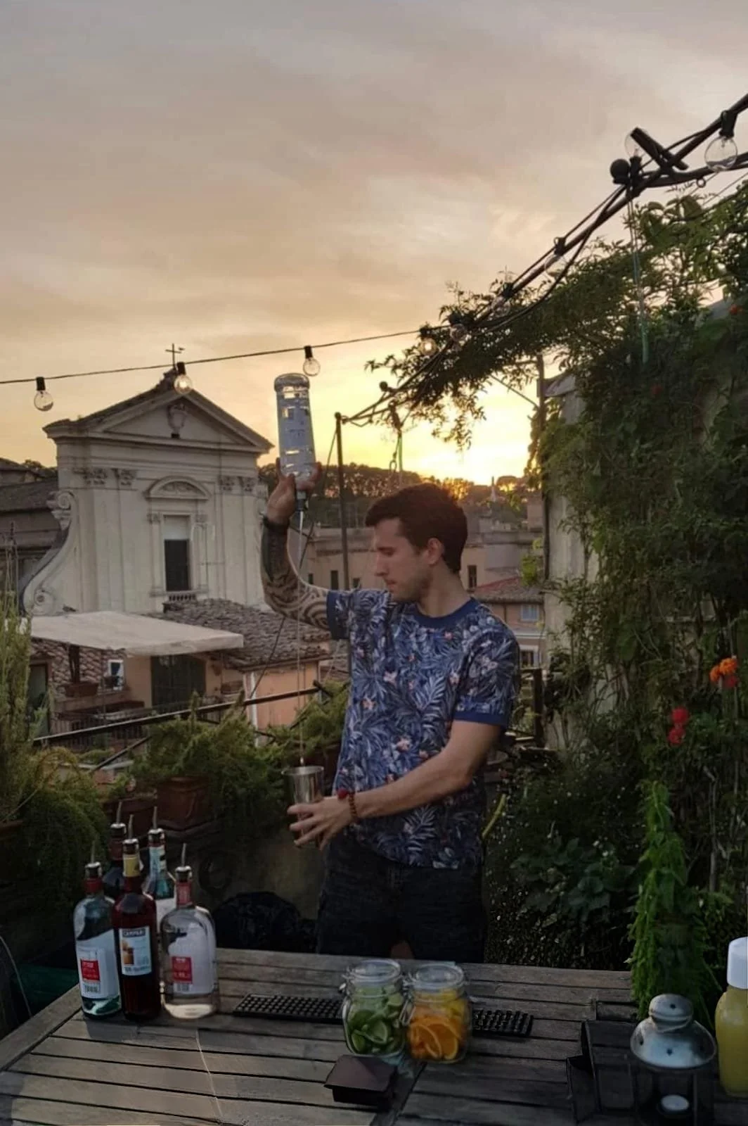 Hombre en una terraza, preparando un cóctel con alcohol, en una puesta de sol, rodeado de plantas y botellas, con un decorativo jardín y cielo nublado.