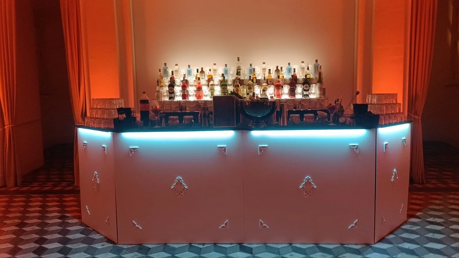 Bar decorativo con botellas y vasos de alcohol, iluminación suave y paredes con cortinas doradas.