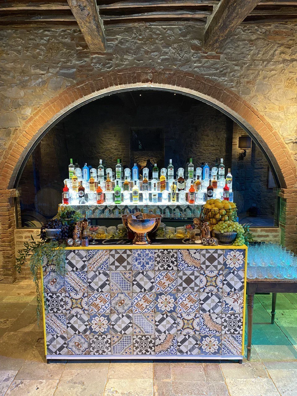 Bar con botellas de licor y decoraciones de frutas y uvas en un ambiente con paredes de piedra y arco de ladrillo.