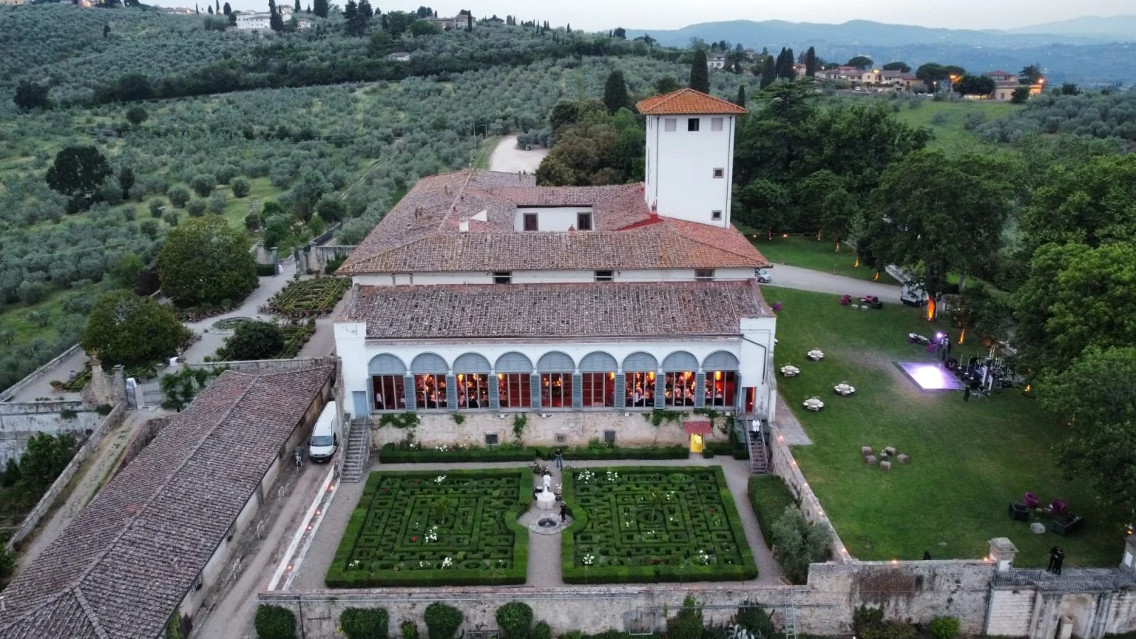 Vista aérea de una posada o villa en un paisaje campestre con jardines, árboles y colinas verdes, al atardecer.
