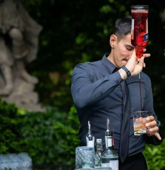 Un hombre con camisa azul oscuro sirviendo un cóctel en un jardín, usando un recipiente grande con líquido oscuro y un vaso en la mano izquierda.
