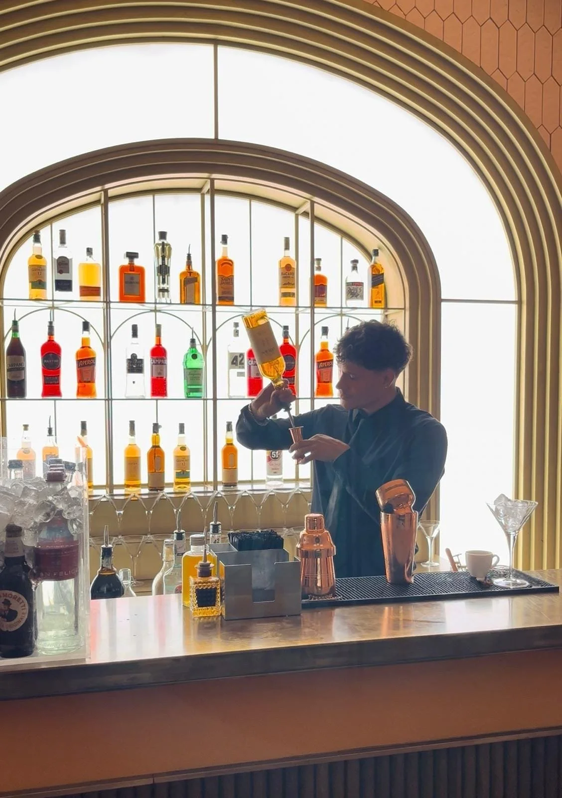 Barman preparando un cóctel en un bar con estantería de botellas de alcohol y gran ventana con luz natural de fondo.
