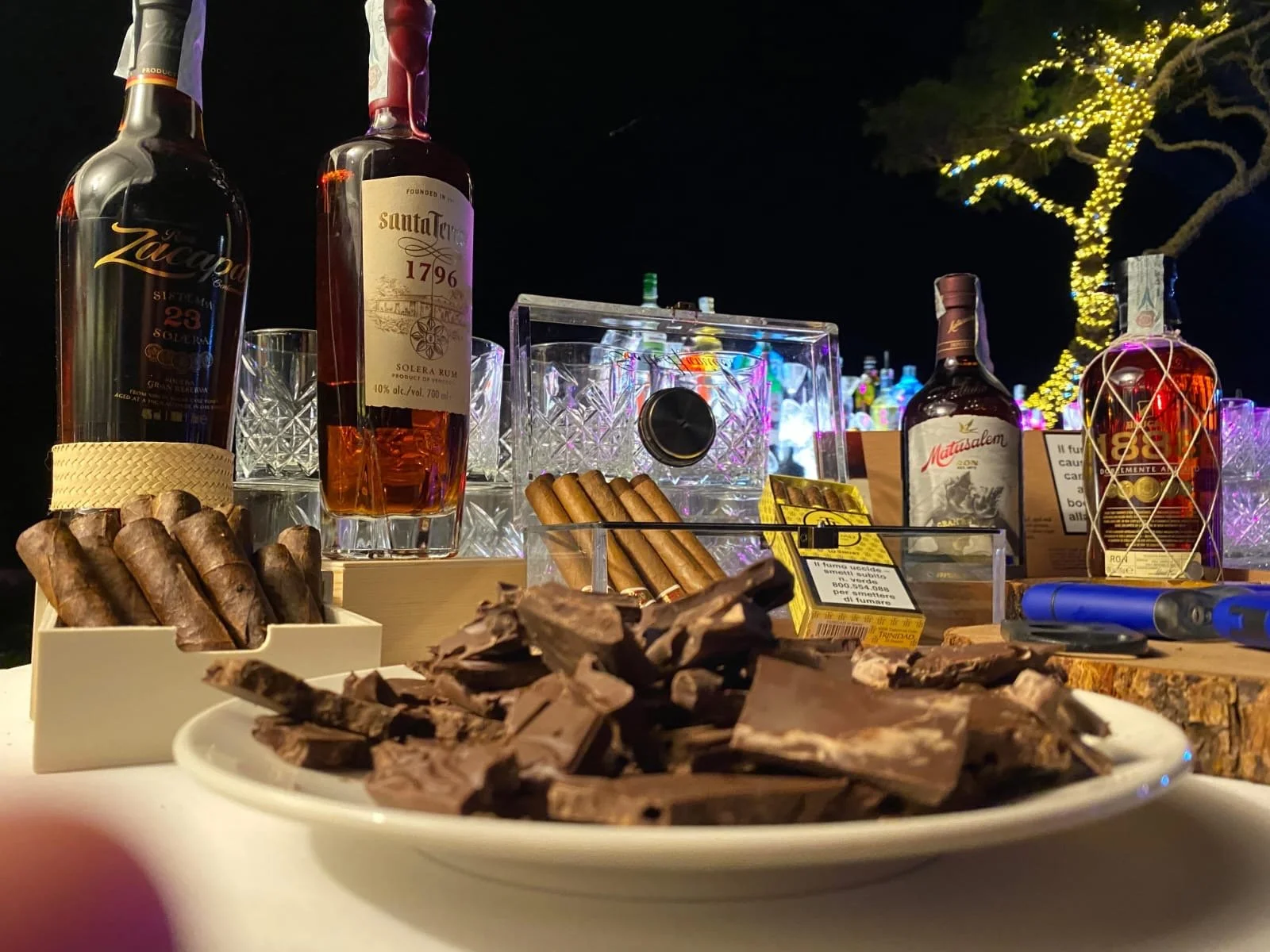 Barra con botellas de licor, cigarros, chocolates y un plato con chocolate picado, decoraciones de luces y vasos en el fondo.