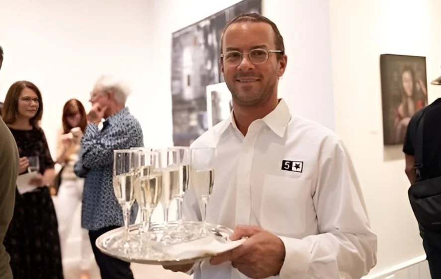 A smiling event server in a white shirt holding a tray of champagne glasses at an art gallery event.