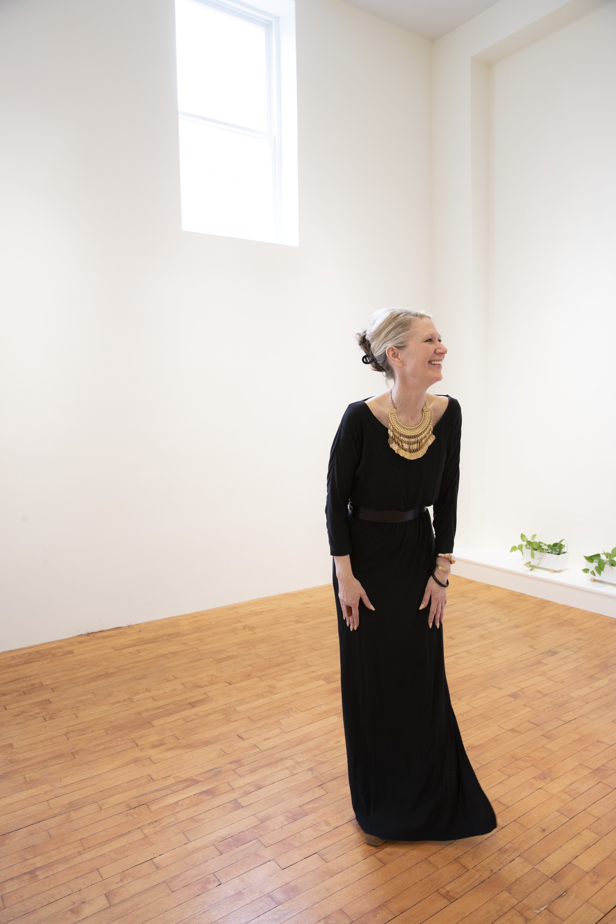 An elderly woman with gray hair tied back, wearing a long black dress and a large gold necklace, smiling and standing in a minimalistic room with wooden flooring, white walls, a window, and some potted plants.