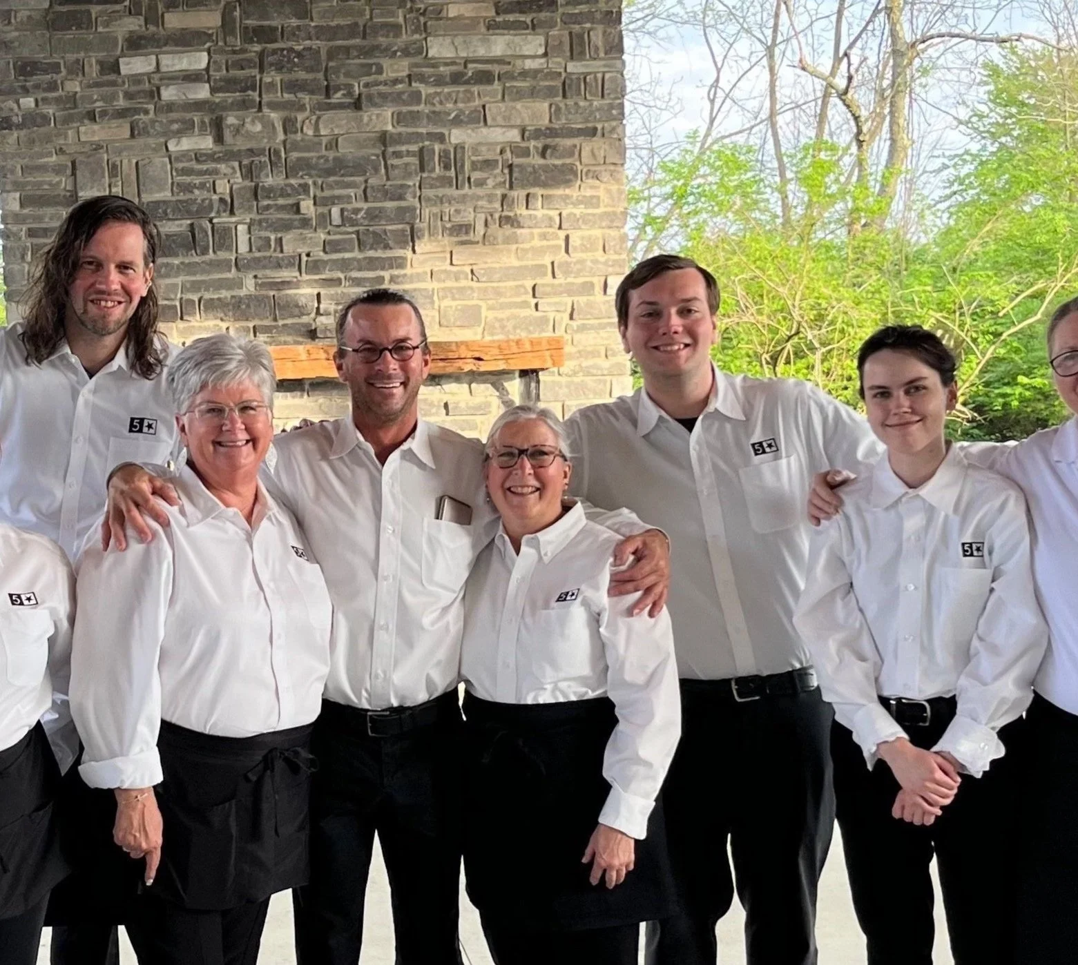 Group of eight event servers wearing matching white shirts with a logo, smiling, standing close together outside with a brick wall and trees in the background.