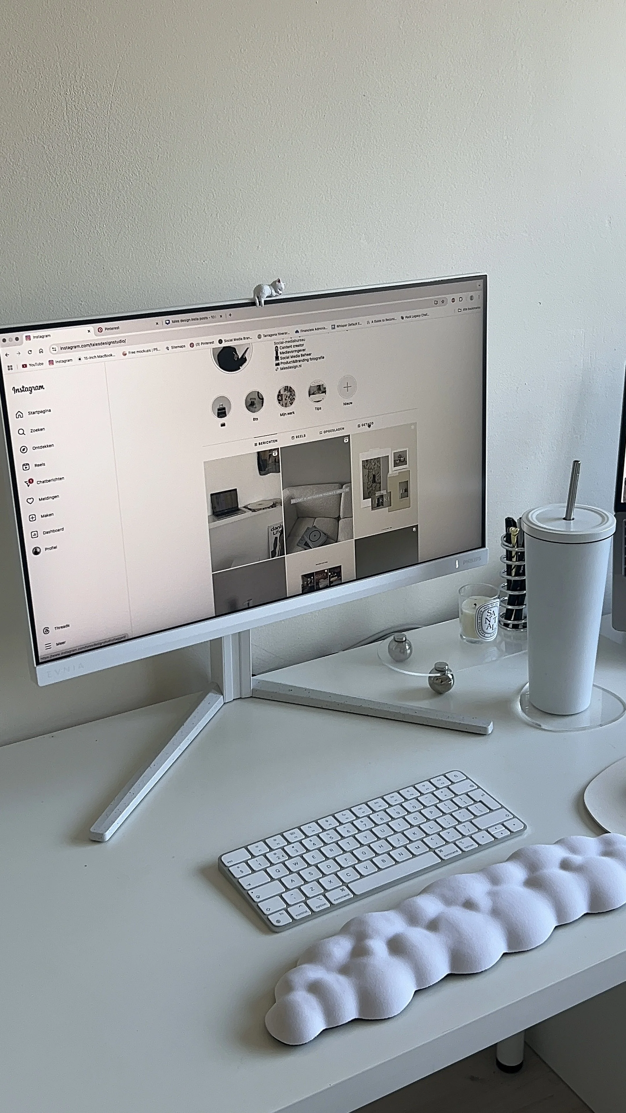 A white desk with a curved foam desk mat, a wireless keyboard, a small glass candle, a large water tumbler with a straw, and a computer monitor showing an Instagram page. There are decorative objects on the desk and a white wall behind.