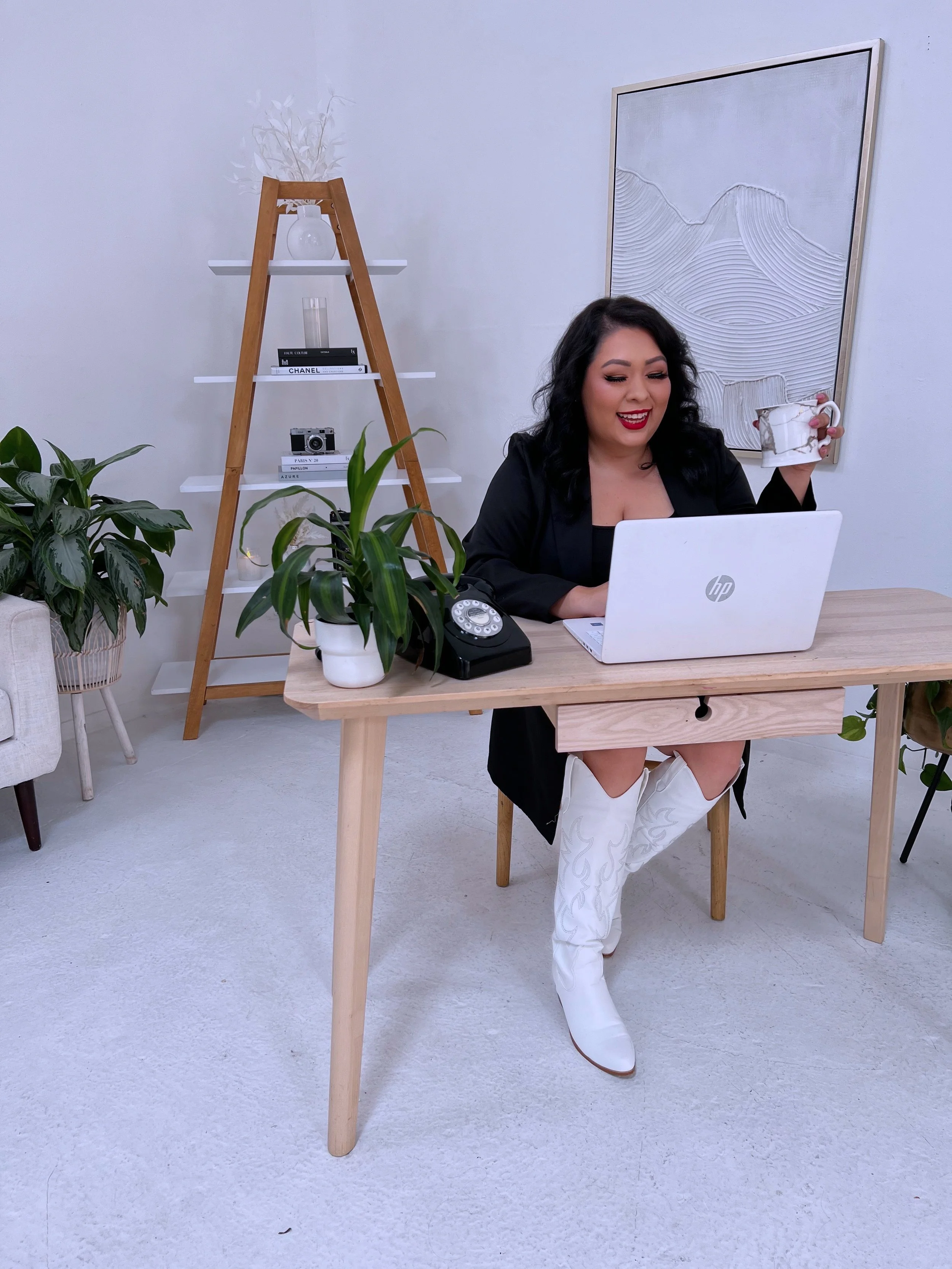 A woman with black curly hair sitting at a wooden desk, holding a ceramic mug with a marble pattern, smiling, in a room with a white wall, a ladder-style shelf with books and decorative items, and plants.