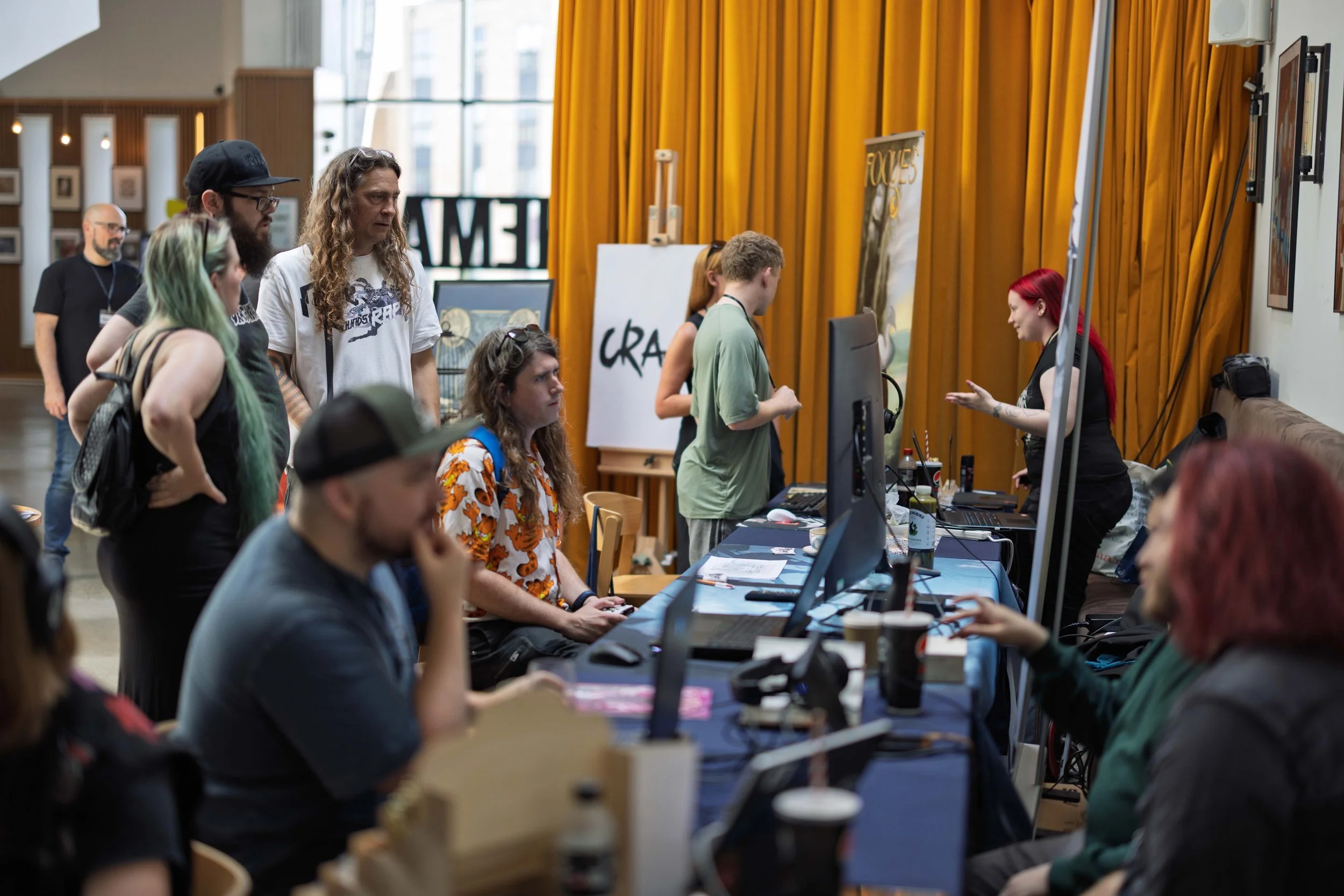 People standing in line at a registration or check-in desk at an event, with a woman with red hair talking to a person behind the desk, in a room with yellow curtains and framed artwork on the wall.