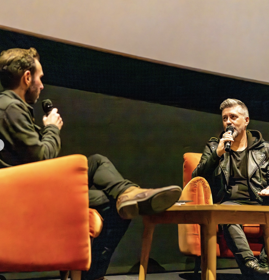 Two men having a conversation on stage, seated in orange chairs, each holding a microphone, in a dimly lit room.