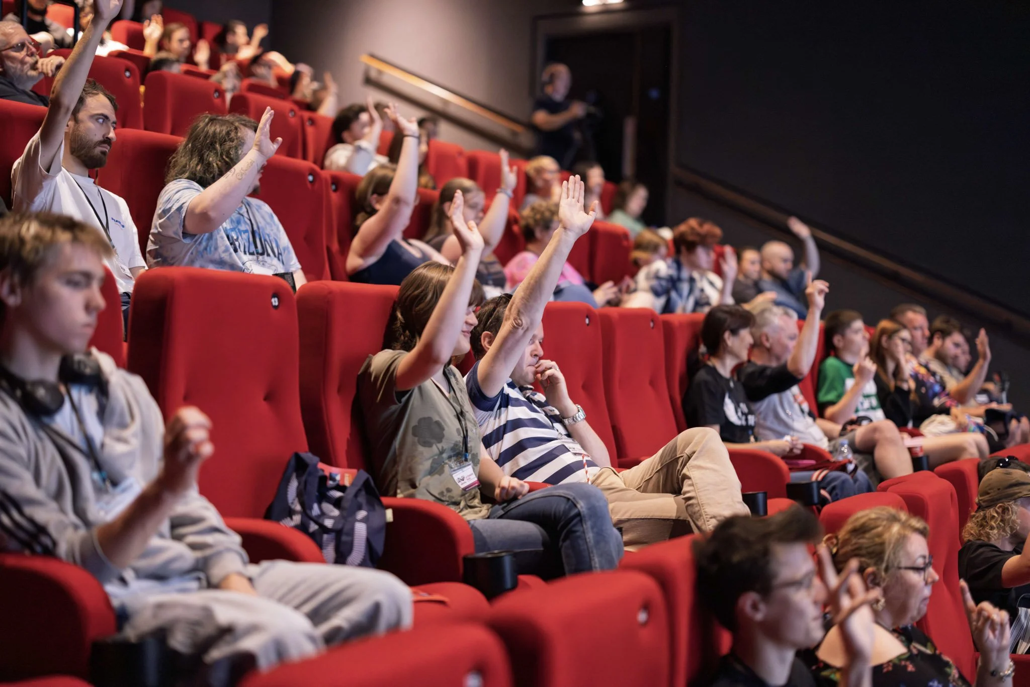 Audience members in a theater-style setting with red seats, many raising their hands, some holding phones, attending an event.