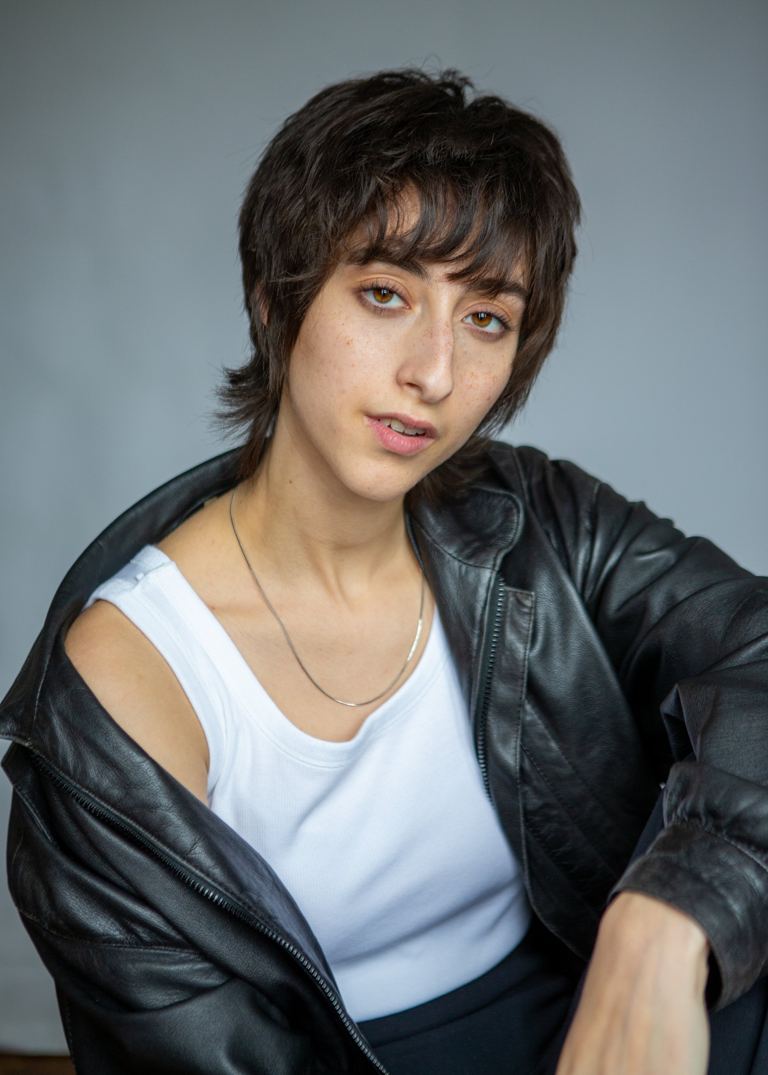 Person with short dark hair wearing a leather jacket and white tank top, looking at the camera.