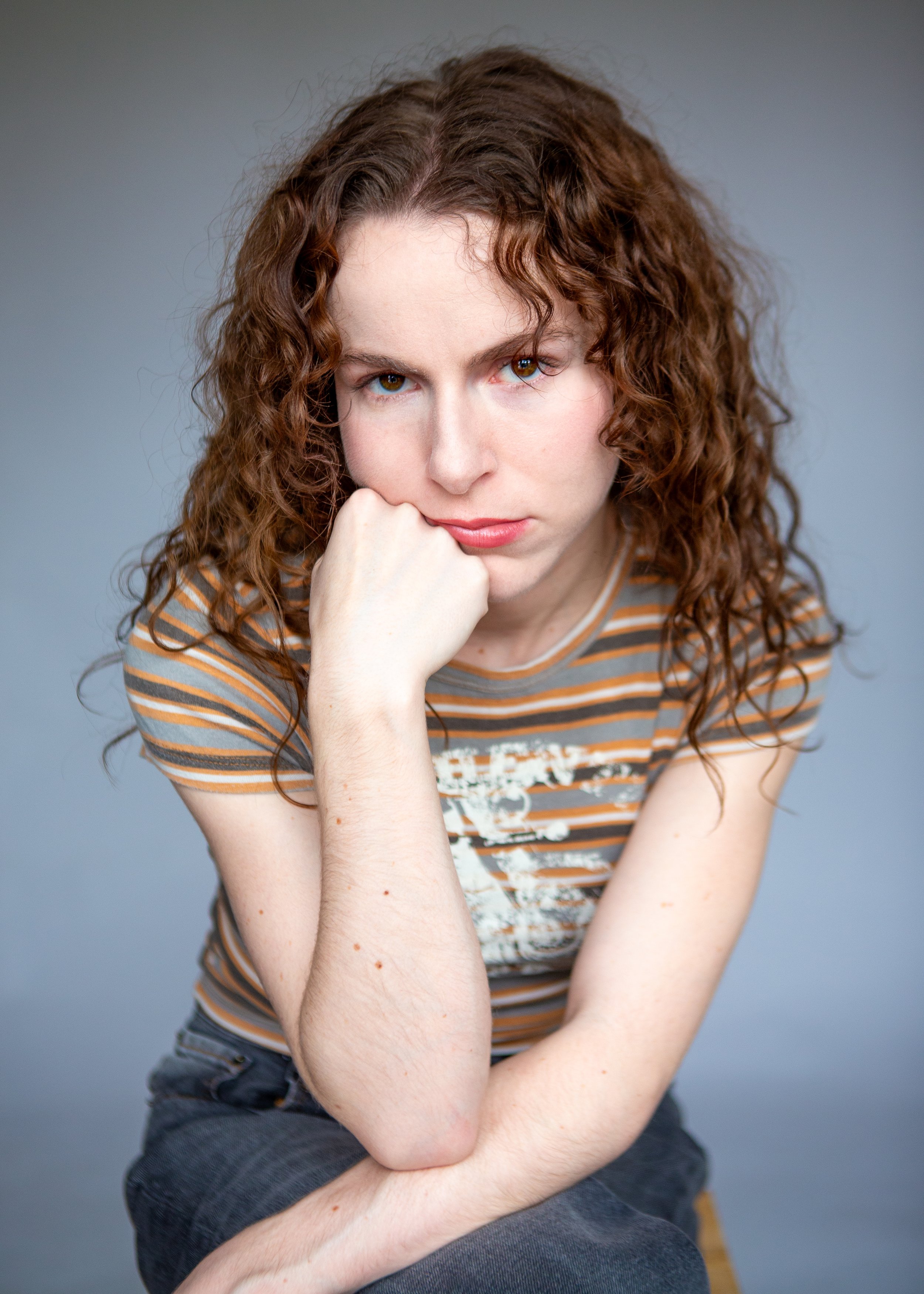 Person with curly hair wearing a striped shirt, resting chin on hand, looking at the camera.