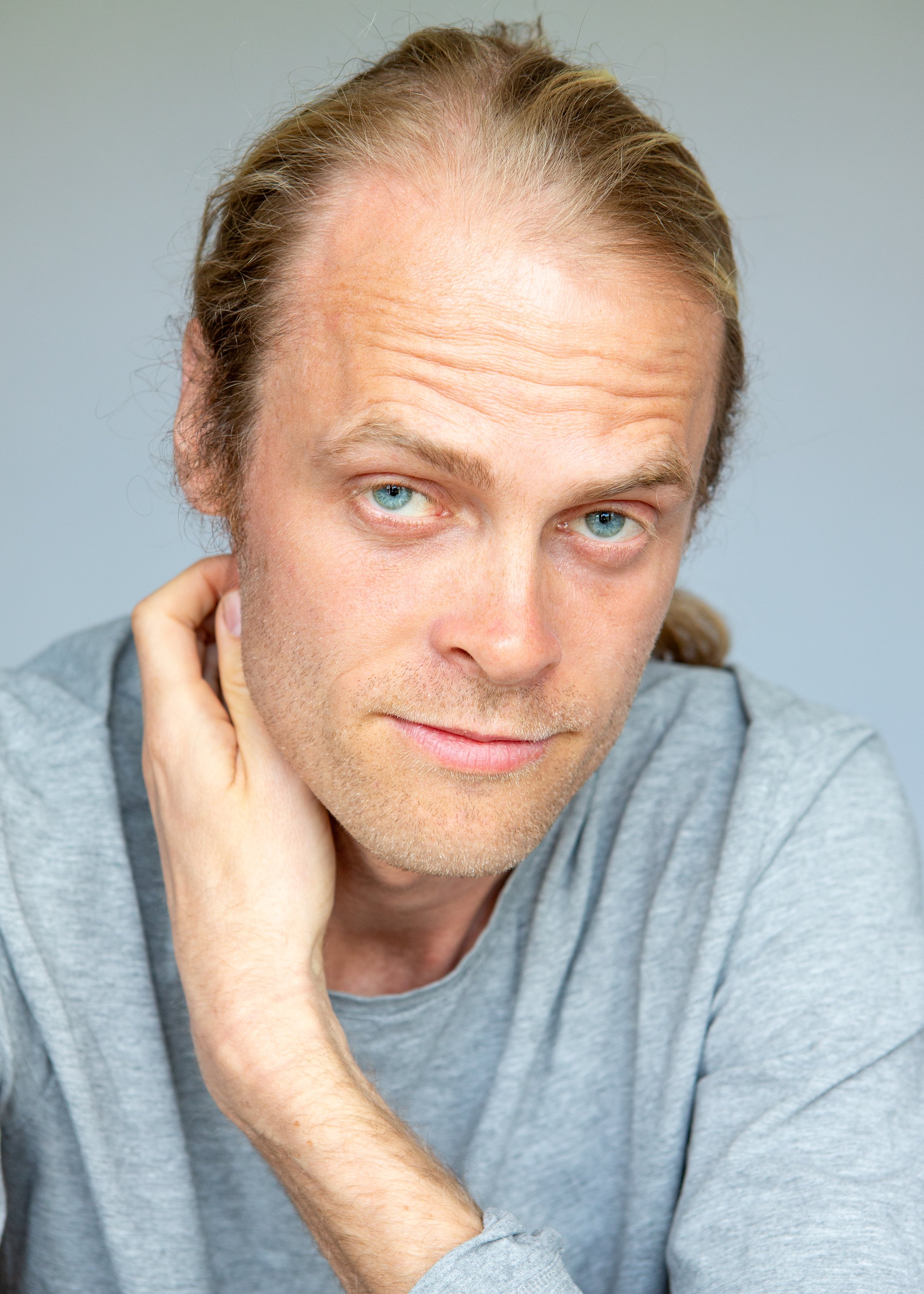 Portrait of a man with light brown hair, wearing a gray shirt, looking at the camera with a hand touching his neck.