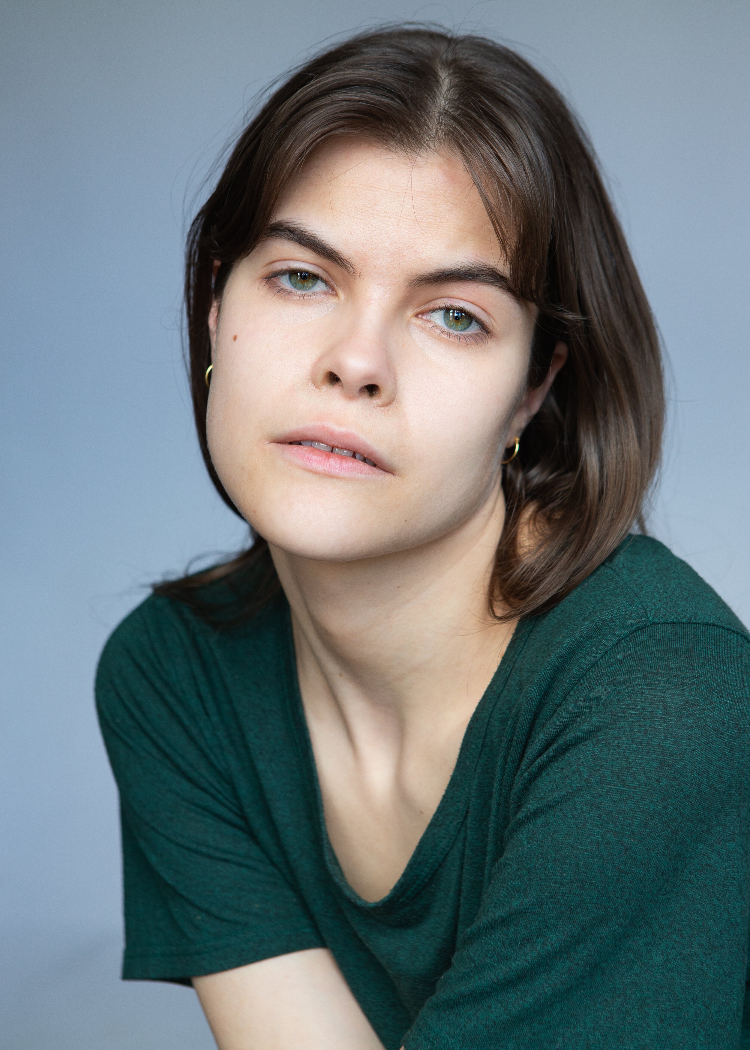 A person with medium-length brown hair and green eyes wearing a dark green shirt, looking at the camera in a neutral expression against a plain background.
