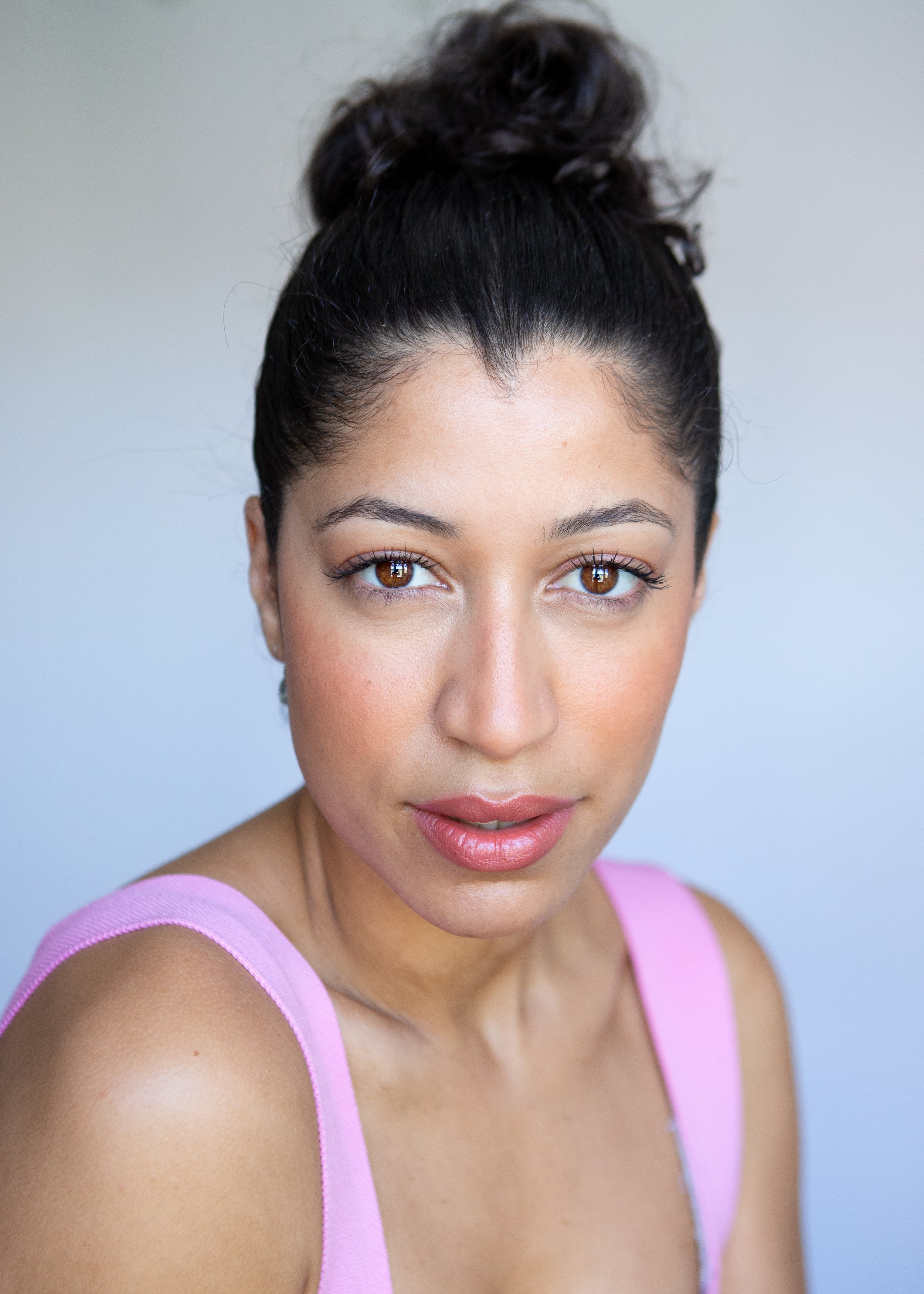 A woman with dark hair in a bun, wearing a pink top, looking directly at the camera.