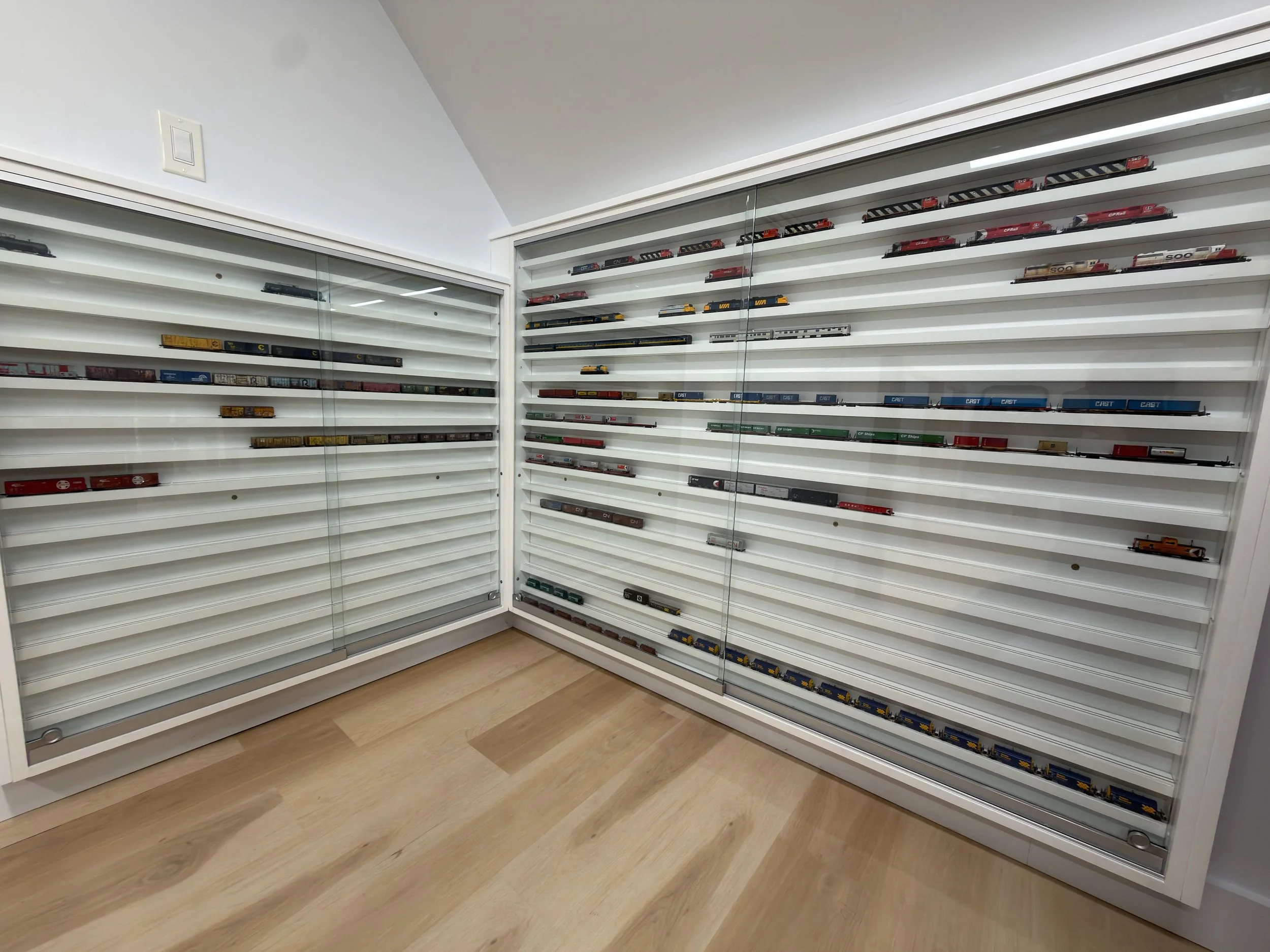 A corner view of white display shelves filled with model train cars in a room with light wooden flooring.