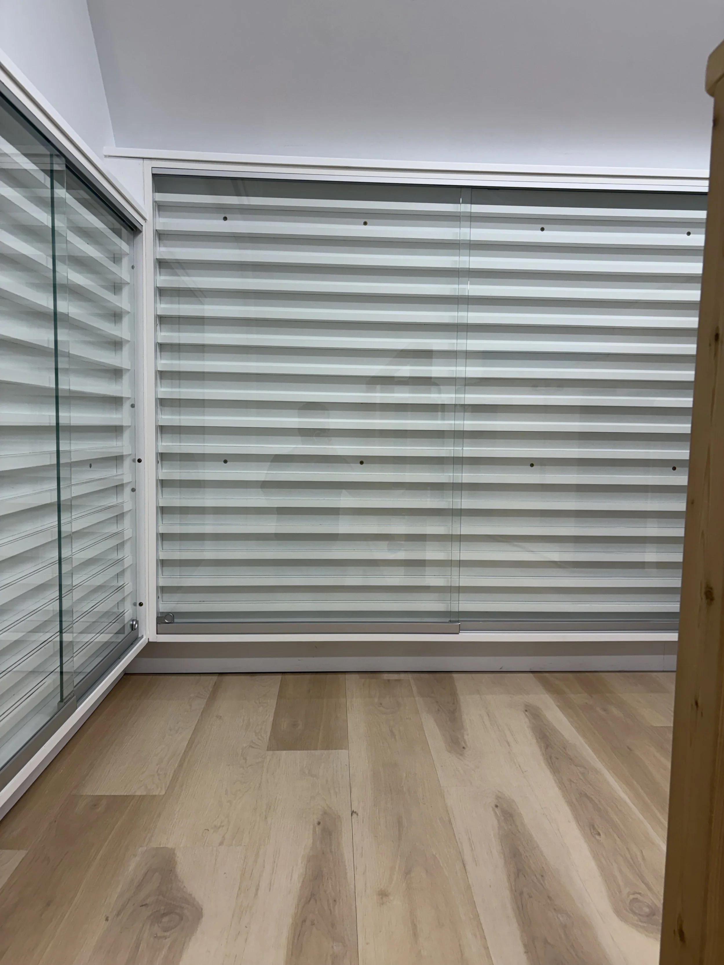 Interior scene with white horizontal slatted window blinds, glass door to the left, and wooden flooring.