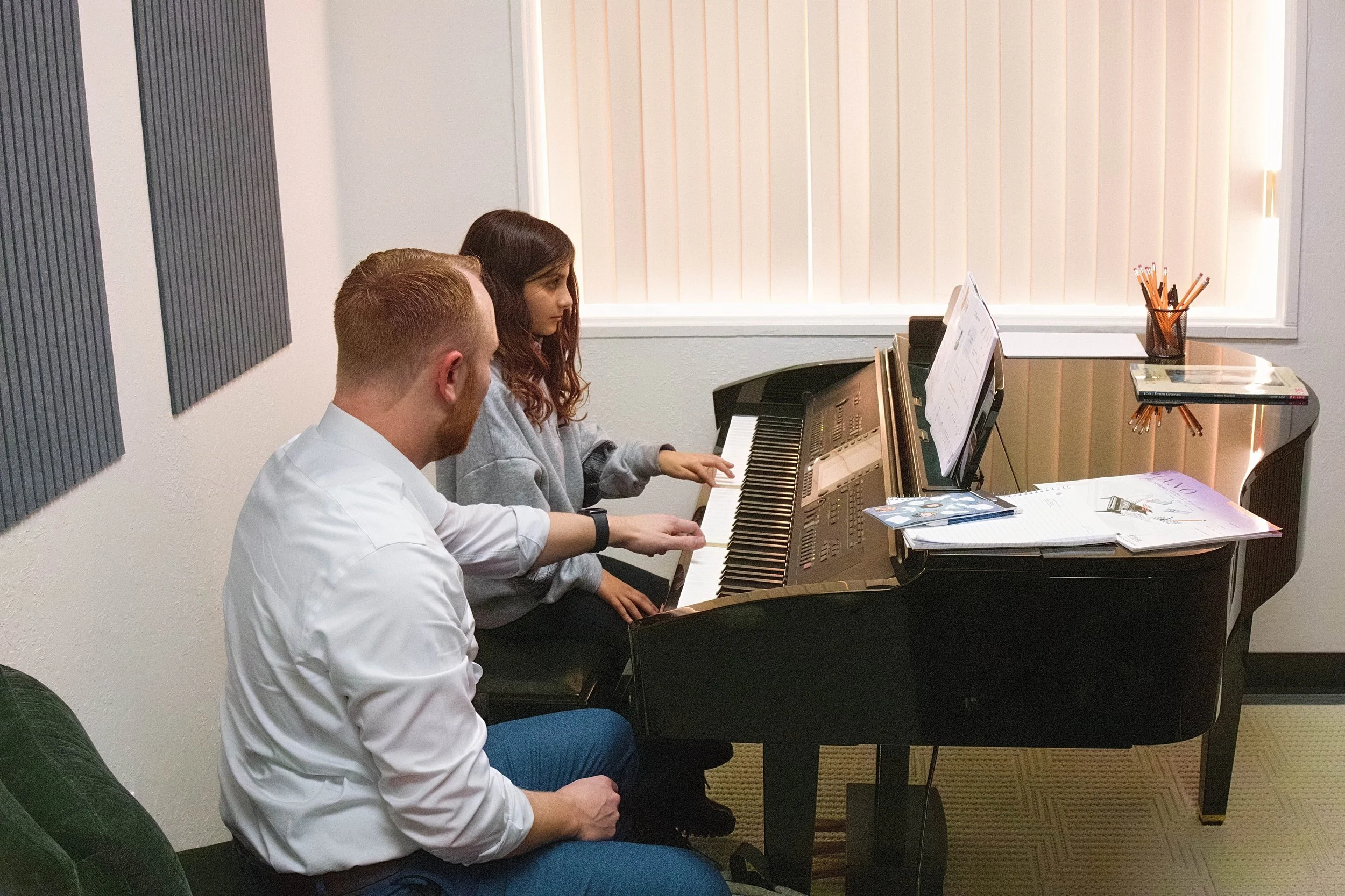 Two young people, a girl with a green baseball cap and a boy with curly red hair, sit closely together reviewing papers, while a woman in the background adjusts a light on a camera setup.