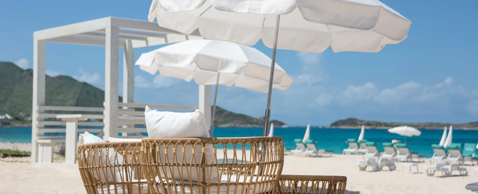 Chaises longues avec parasols blancs sur une plage de sable blanc, avec la mer turquoise et des montagnes en arrière-plan à Orient Bay, Saint-Martin.