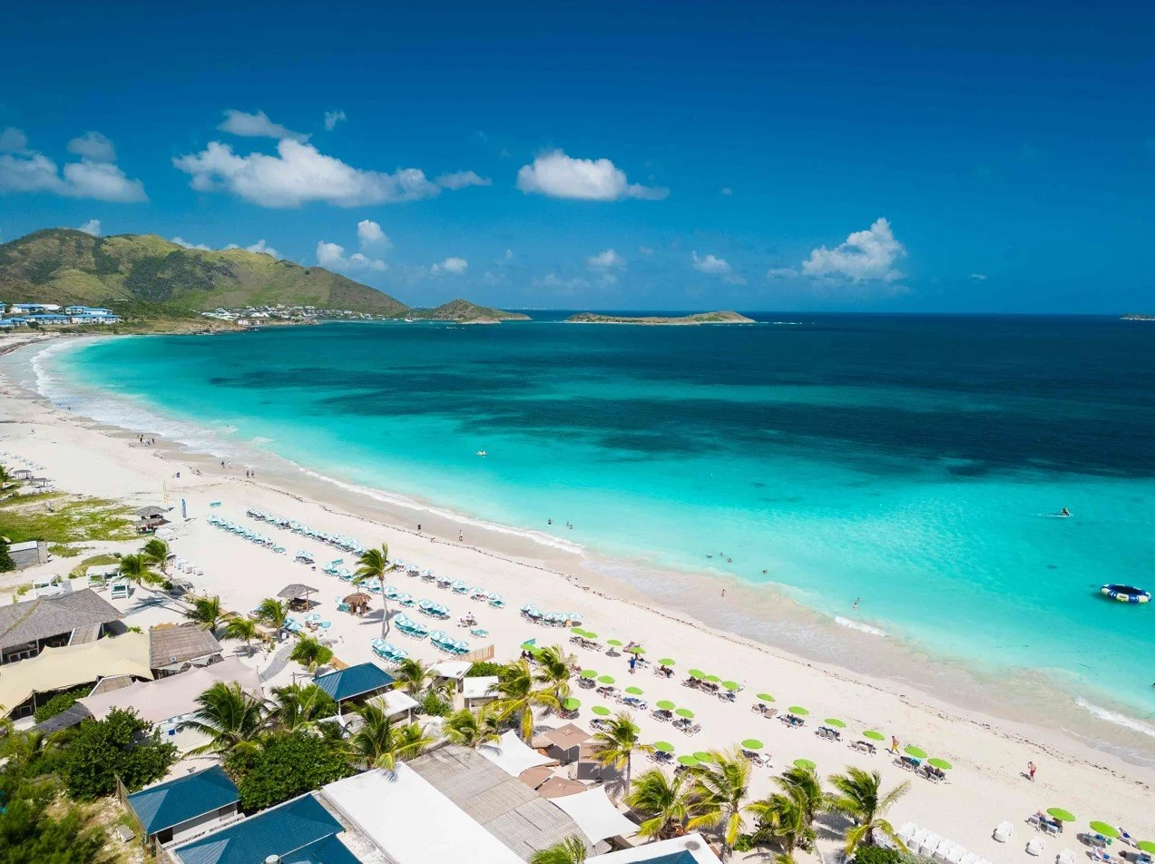Plage de sable blanc avec des parasols verts et des palmiers, eau turquoise, montagnes en arrière-plan, ciel bleu avec quelques nuages à Orient Bay, Saint-Martin.