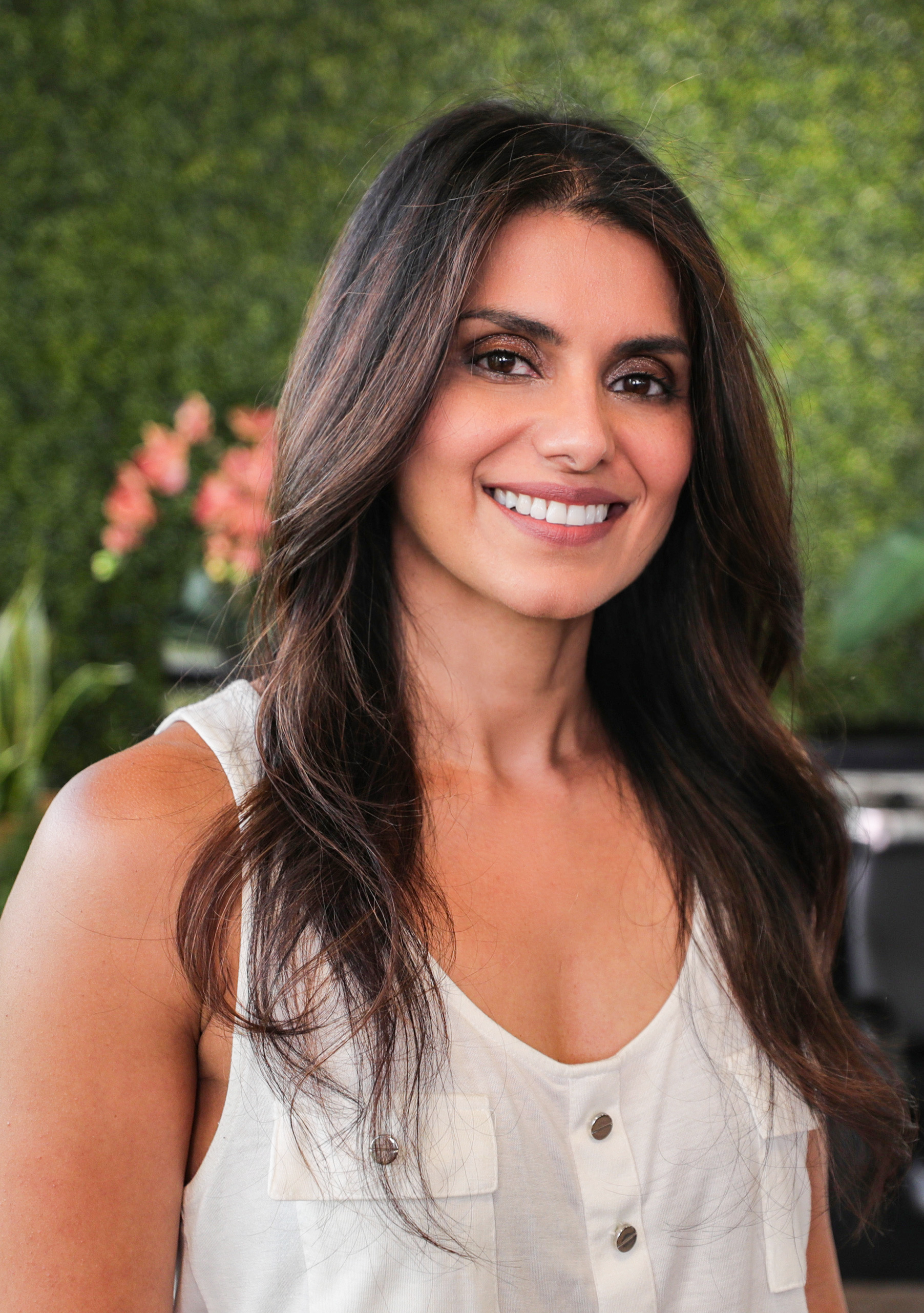 A woman with long brown hair smiling outdoors, with green foliage and pink flowers in the background.