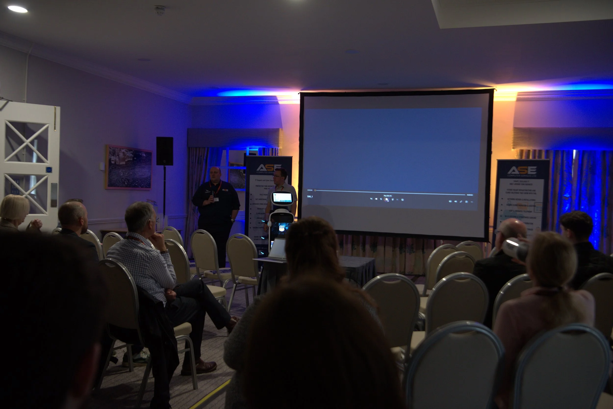 A conference room with people seated facing a large projection screen, with two presenters standing near the screen, and a dimly lit room with blue accent lighting.