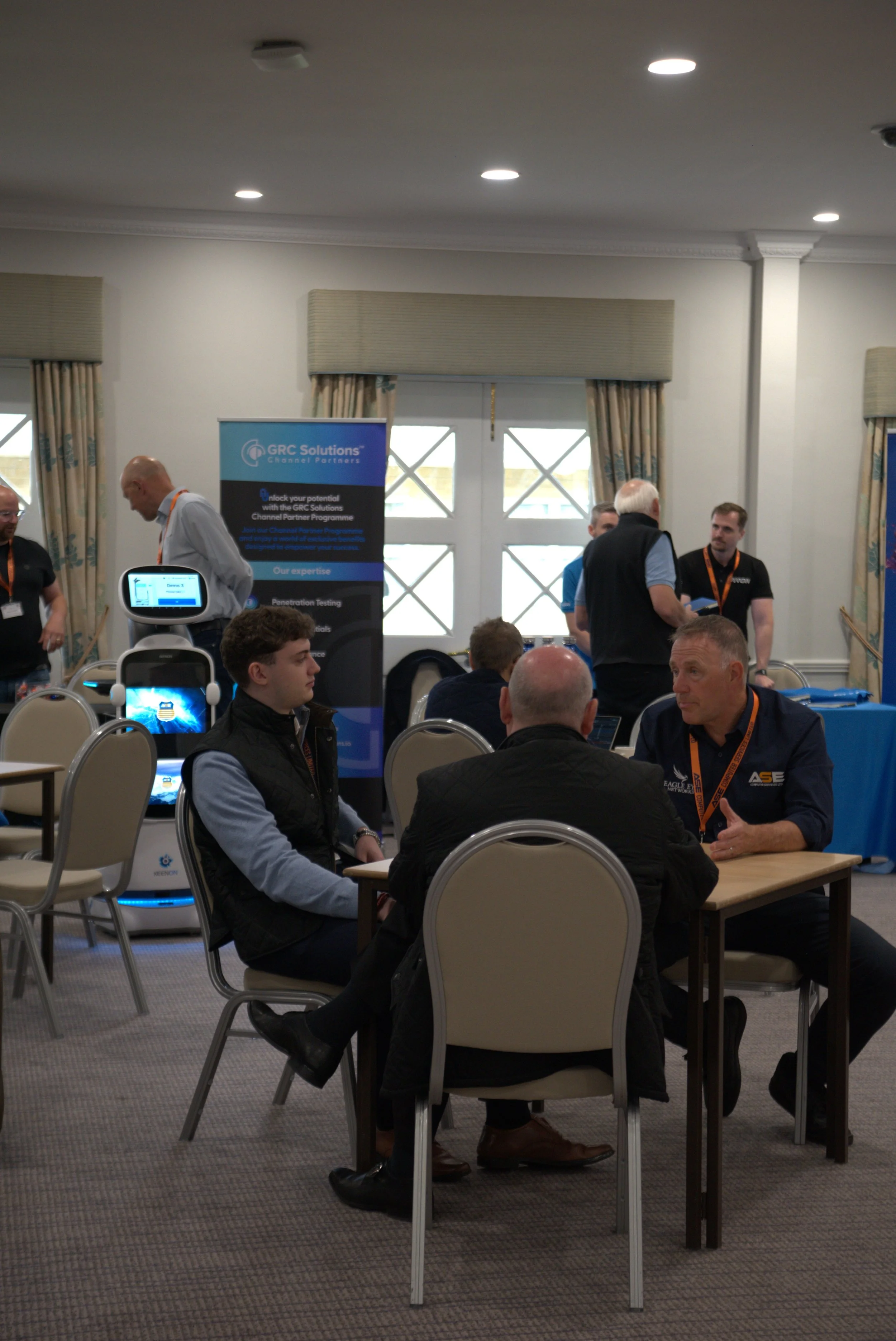 People sitting and talking at a trade show or conference, with banners and display booths in the background.