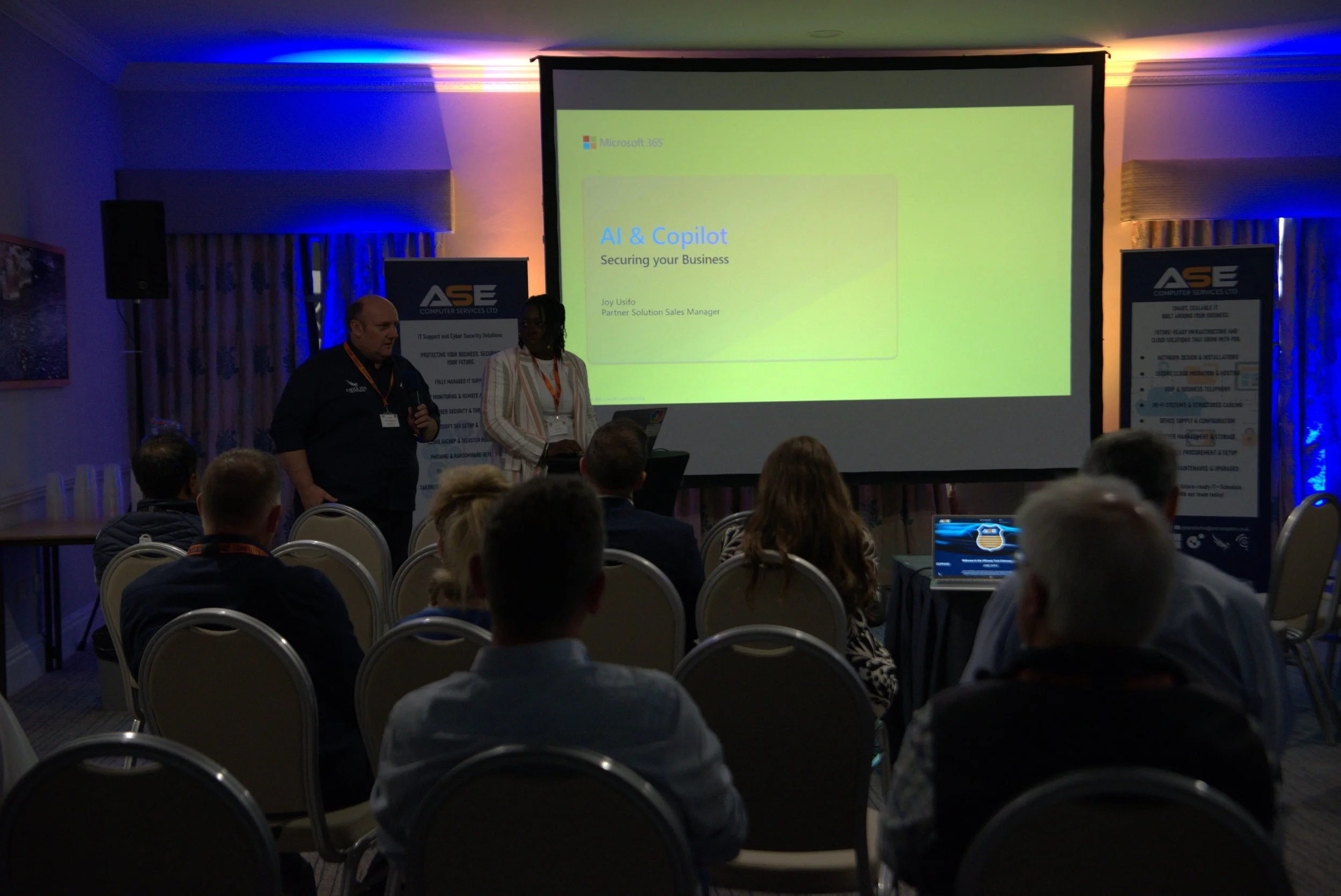 A professional conference room with a presentation screen displaying a slide titled 'AI & Copilot: Securing your Business' by Joy Usifo, with an audience of about ten people seated and two presenters standing near the screen. The room has dim lighting with colorful accents on the walls and contains banners and a laptop on a table.