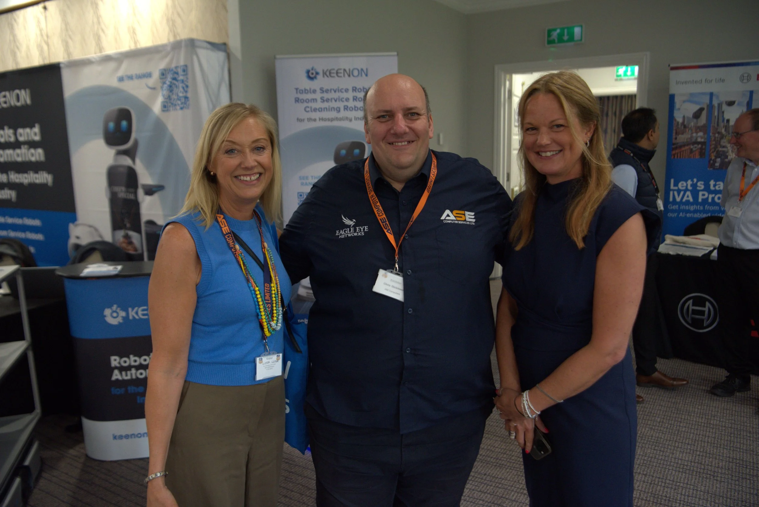 Three people standing together at a conference, smiling at the camera. Two women on either side of a man, all wearing conference badges and branded clothing. Backdrop features banners with technology and robotics branding.